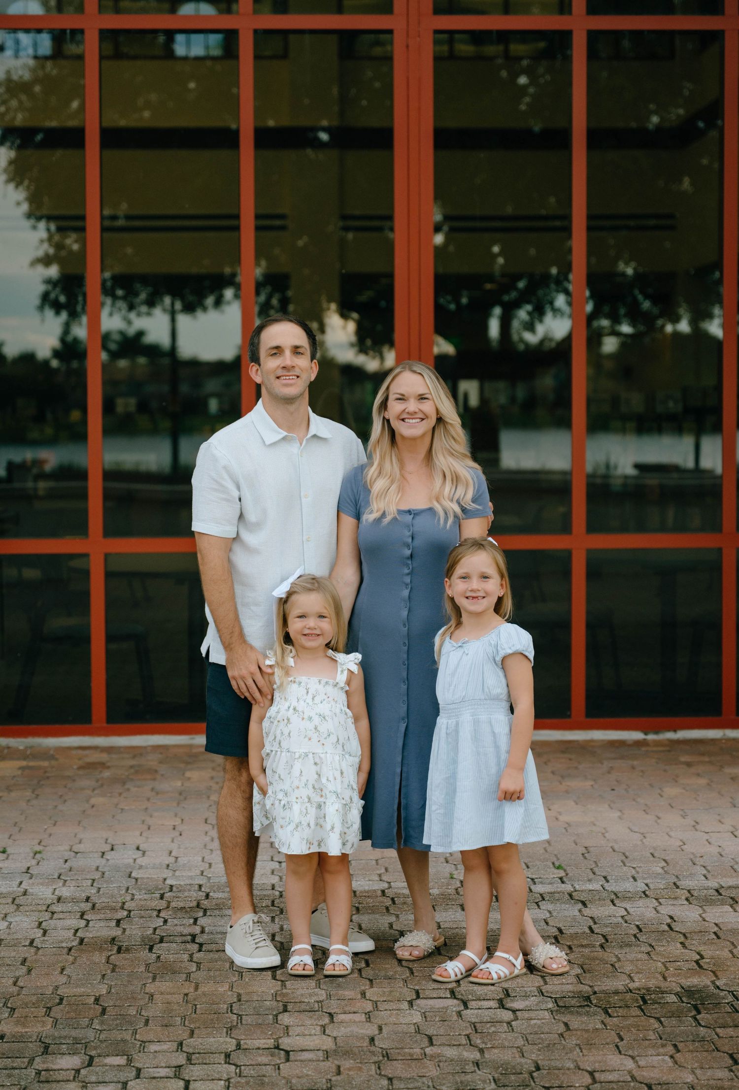 A smiling family of four poses in front of a building with large red-framed windows.