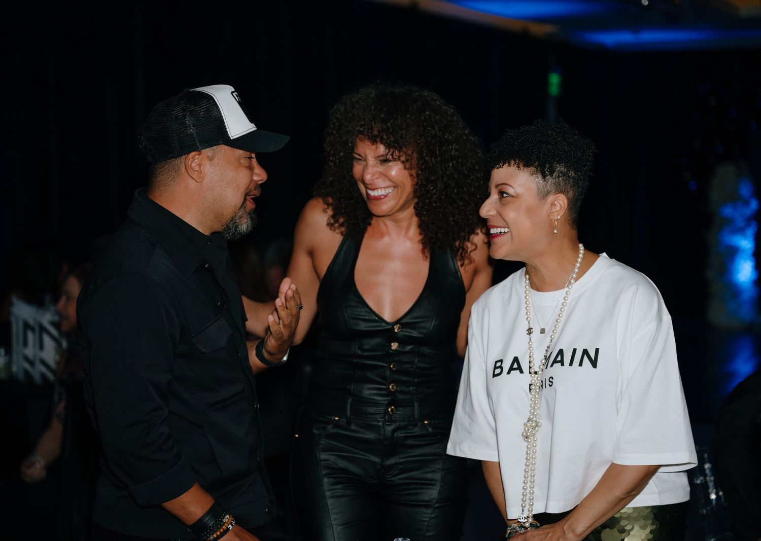 Three friends laugh and socialize together at a nightclub event, one wearing a Balmain t-shirt.