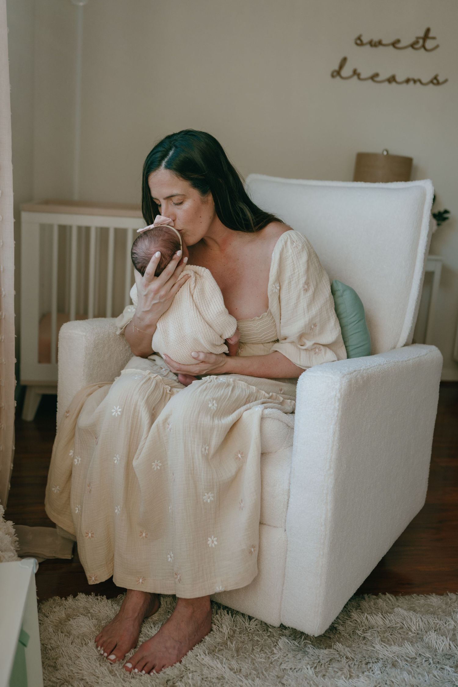 A dark-haired mother in a cream dress kisses her newborn baby while seated in a white nursery chair.