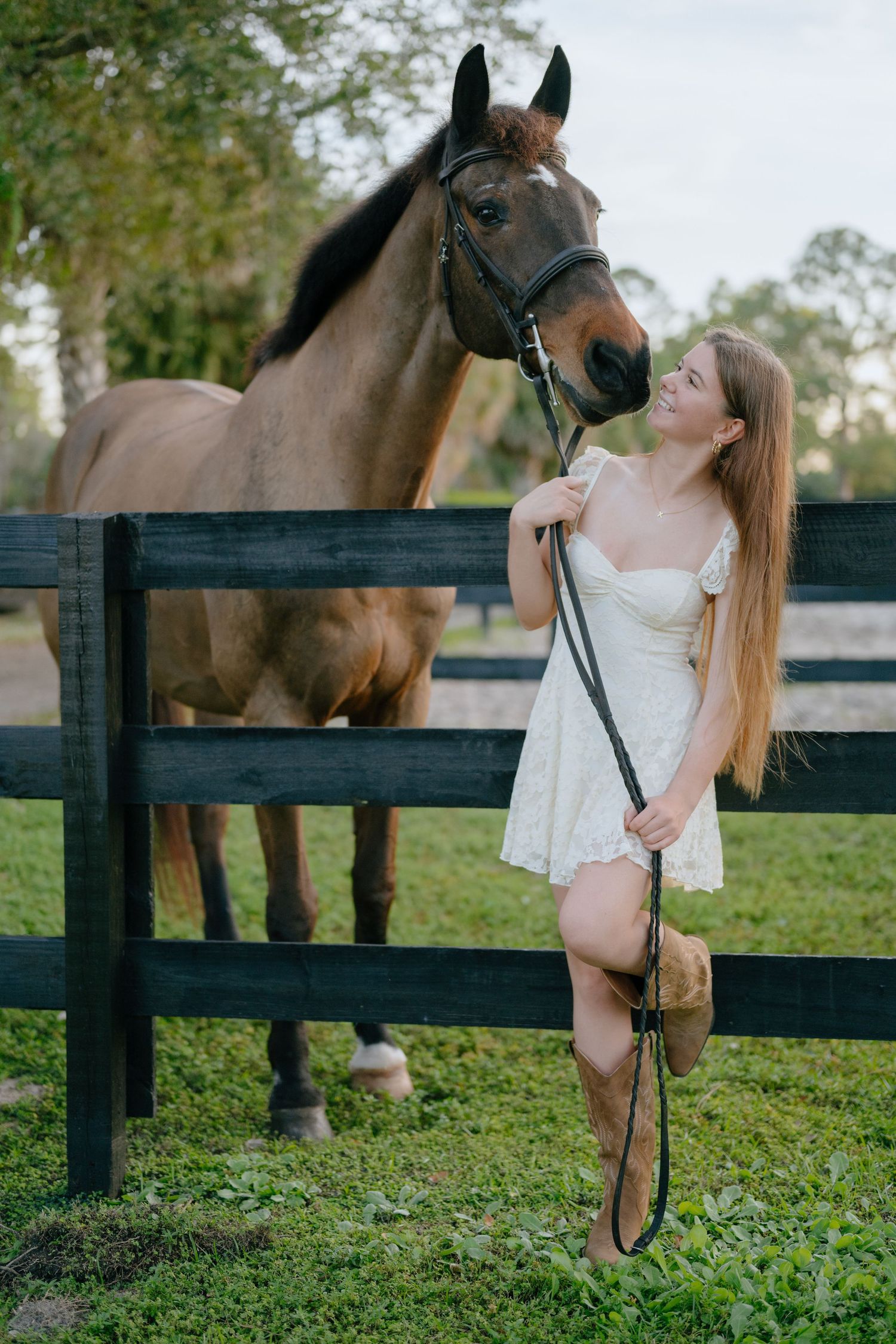 Young woman in white dress smiles at a bay horse while holding reins by a black wooden fence.