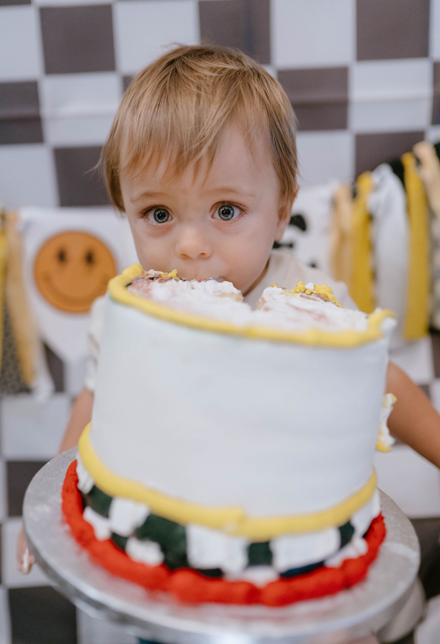 Curious toddler peeks over a decorated first birthday smash cake with checkered and colorful frosting.