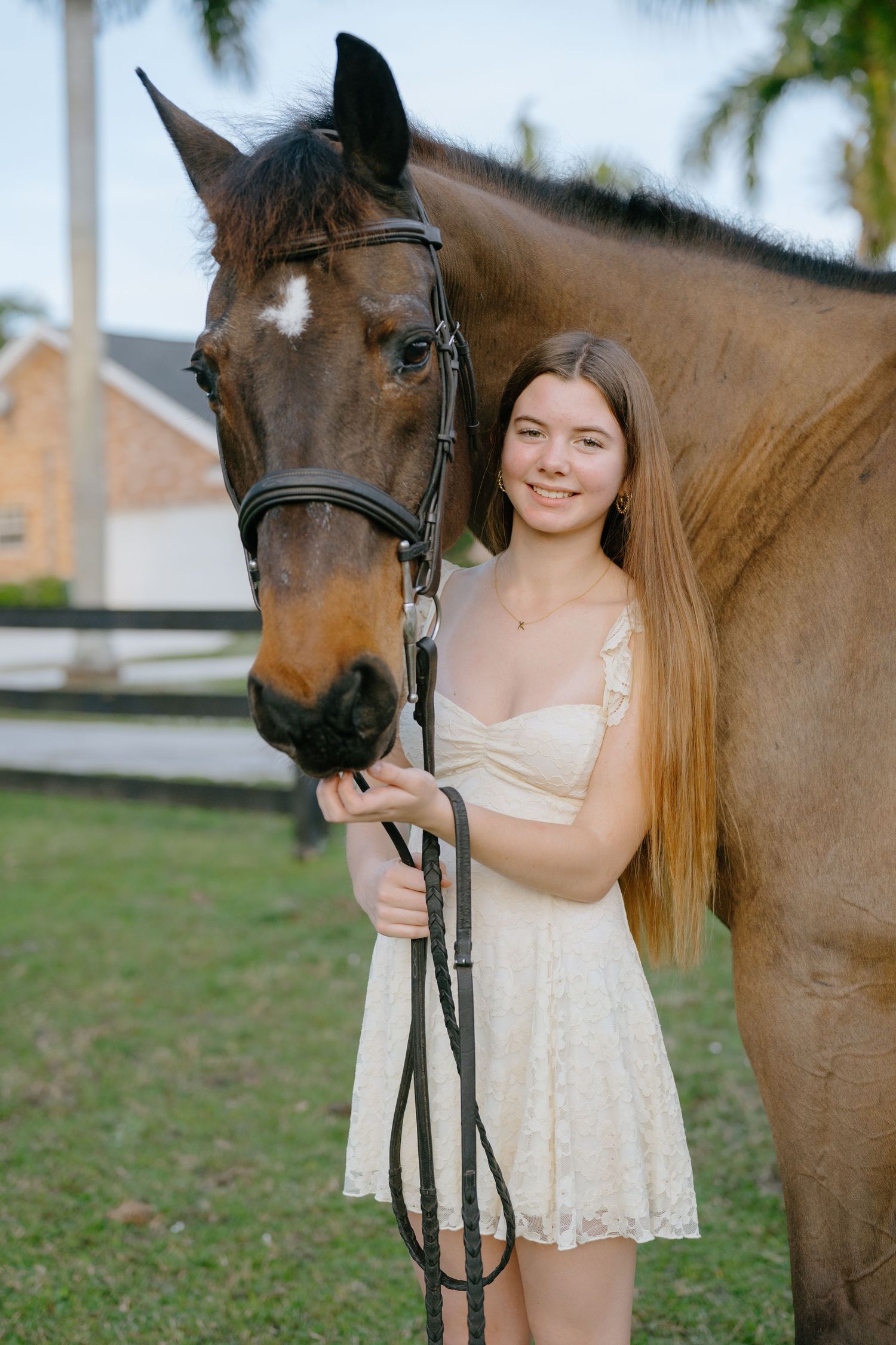 A smiling teenage girl in a cream dress stands proudly beside a bay horse in a sunny paddock.