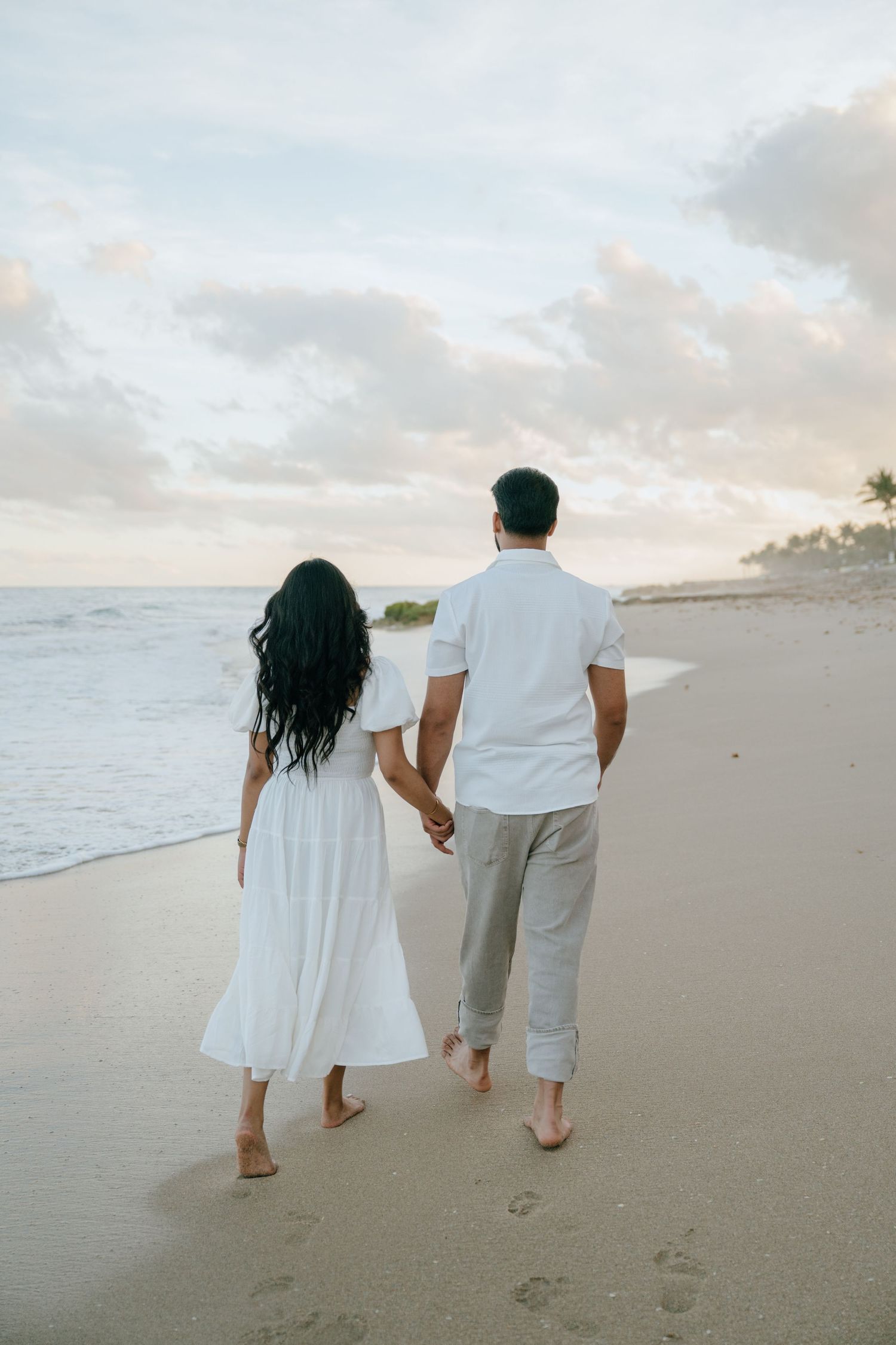 Couple walking hand in hand on a tropical beach at sunset in white outfits, leaving footprints.