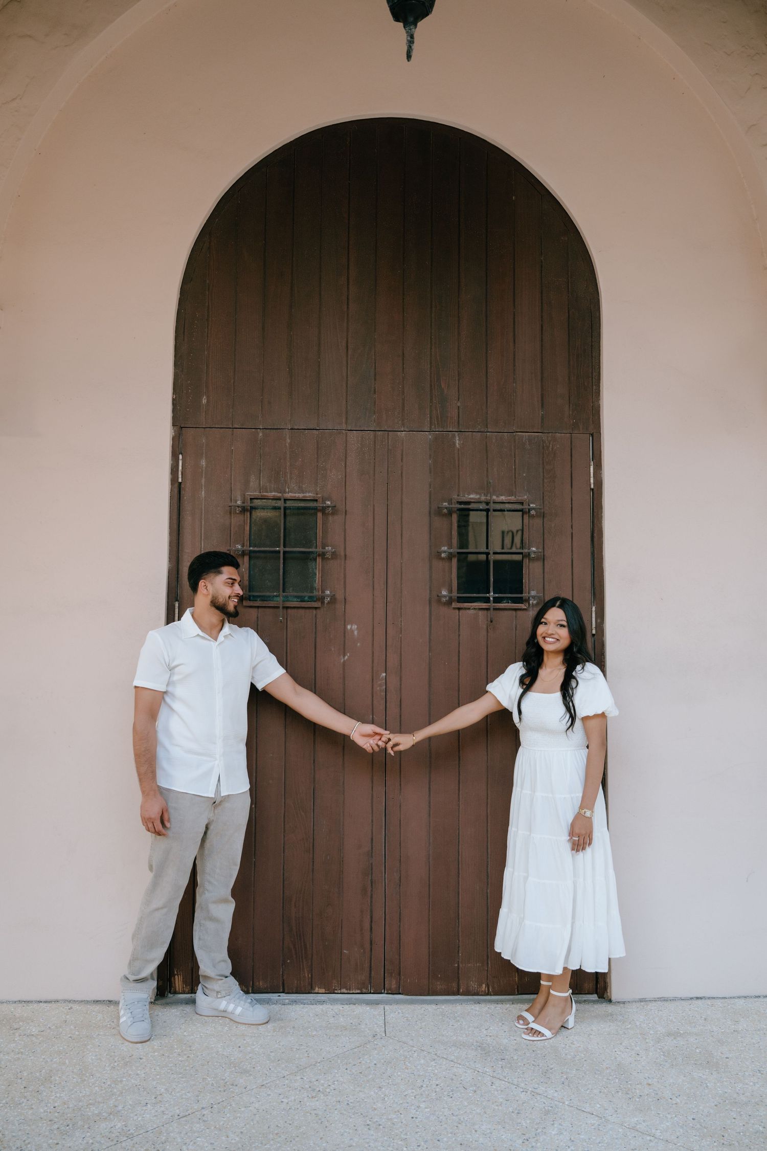 Couple holding hands in front of ornate wooden arched door, man in white shirt and woman in white dress.