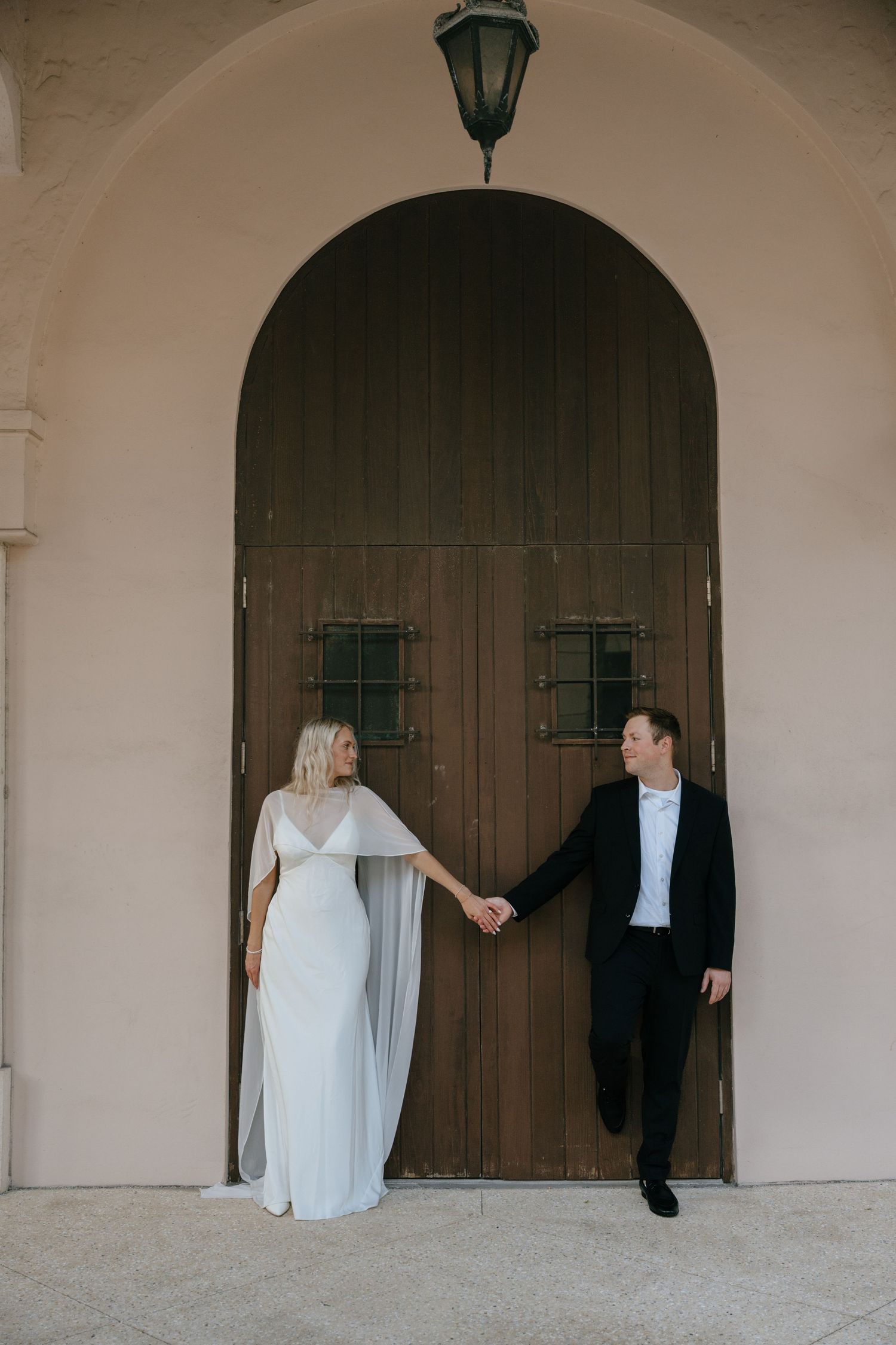 Wedding couple holds hands before a grand arched wooden door, bride in elegant white cape dress.