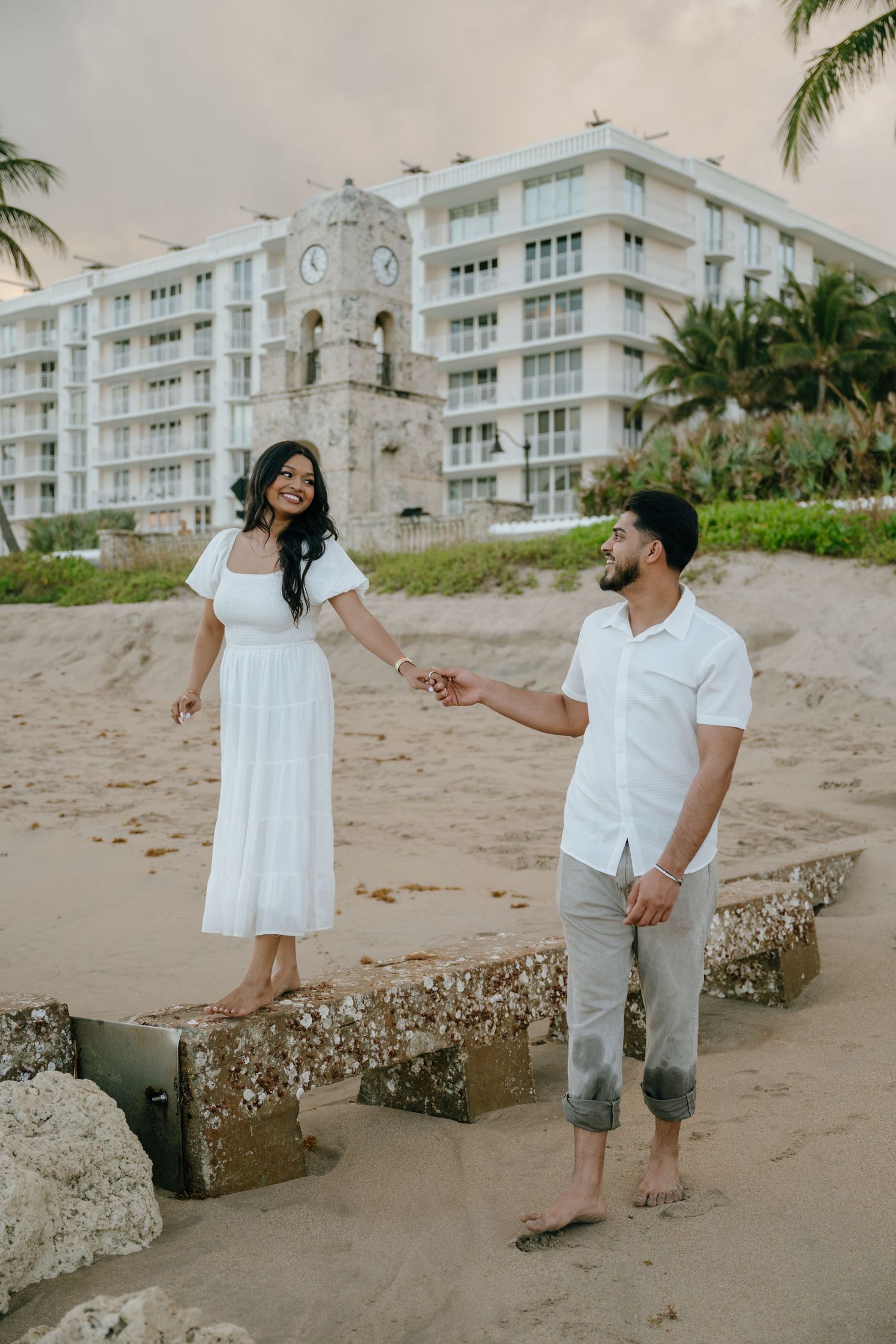 Couple holding hands on sandy beach near historic clock tower, both dressed in white and gray.