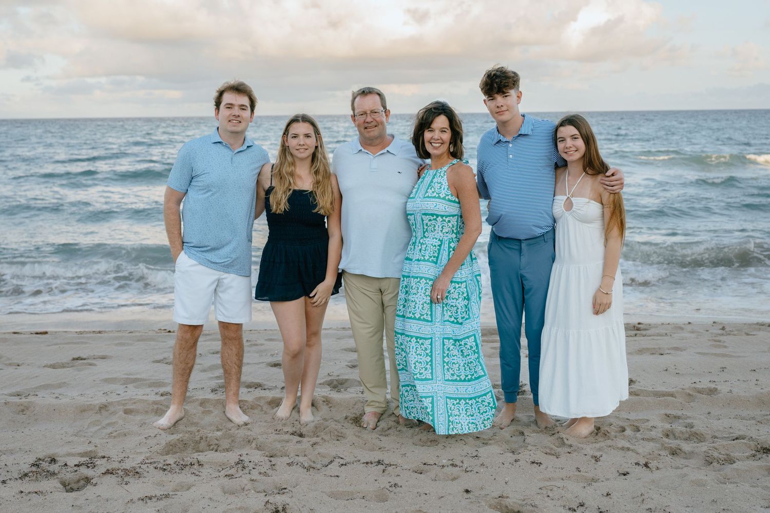 Family of six smiling together on a beach at sunset in coordinated blue and white outfits.