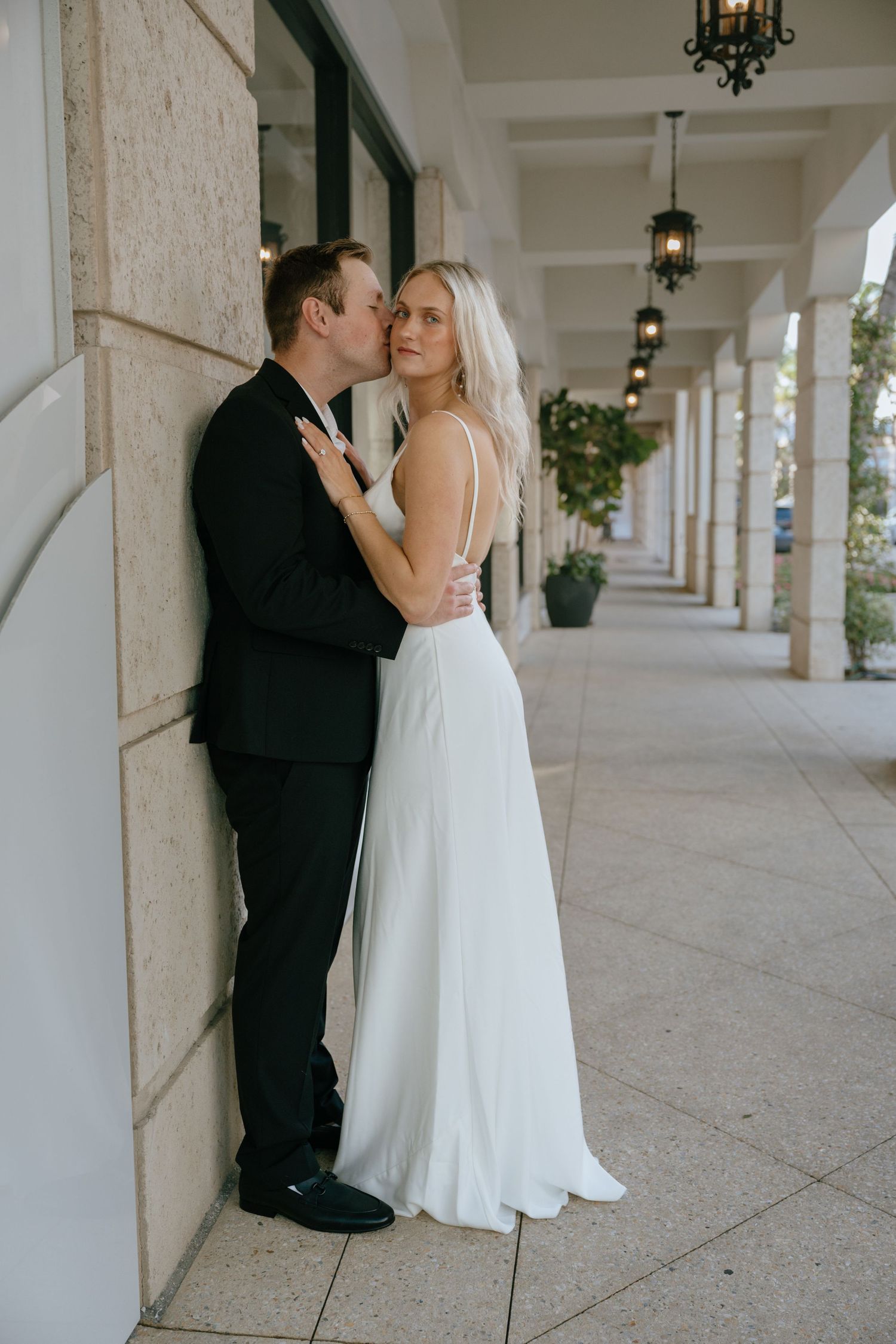 Groom in black suit kissing blonde bride in backless white gown against stone pillar in corridor.