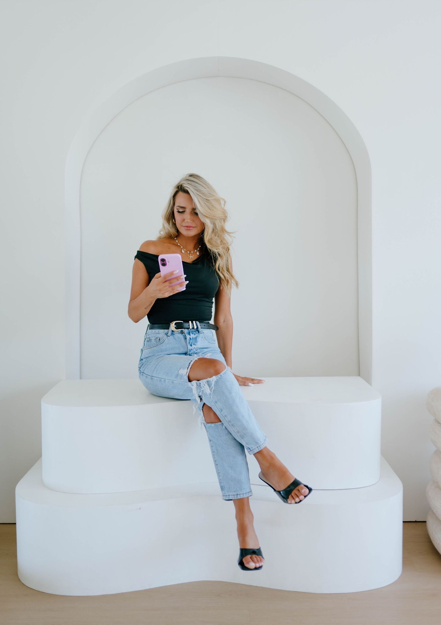 Stylish blonde woman in black off-shoulder top and ripped jeans looking at a pink phone in an arch alcove.