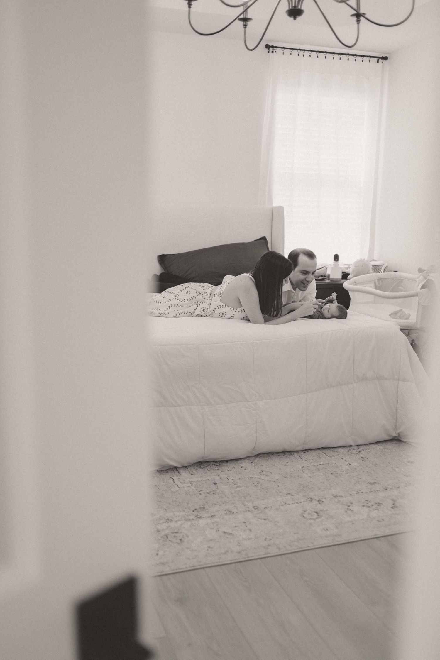 Black and white image of couple on bed with newborn, viewed through doorway.