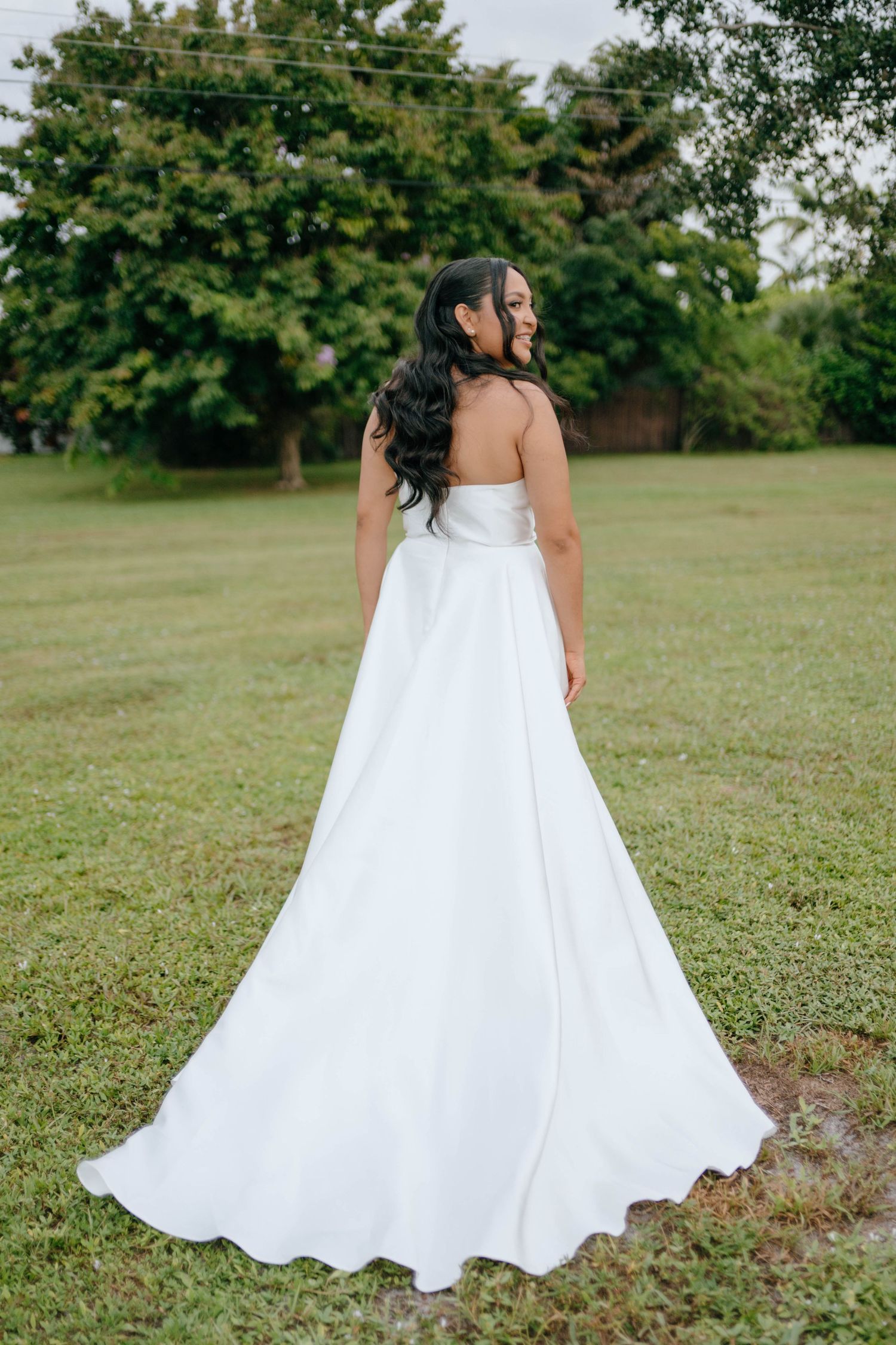 Elegant bride in strapless white ball gown with long train poses outdoors in green garden setting.