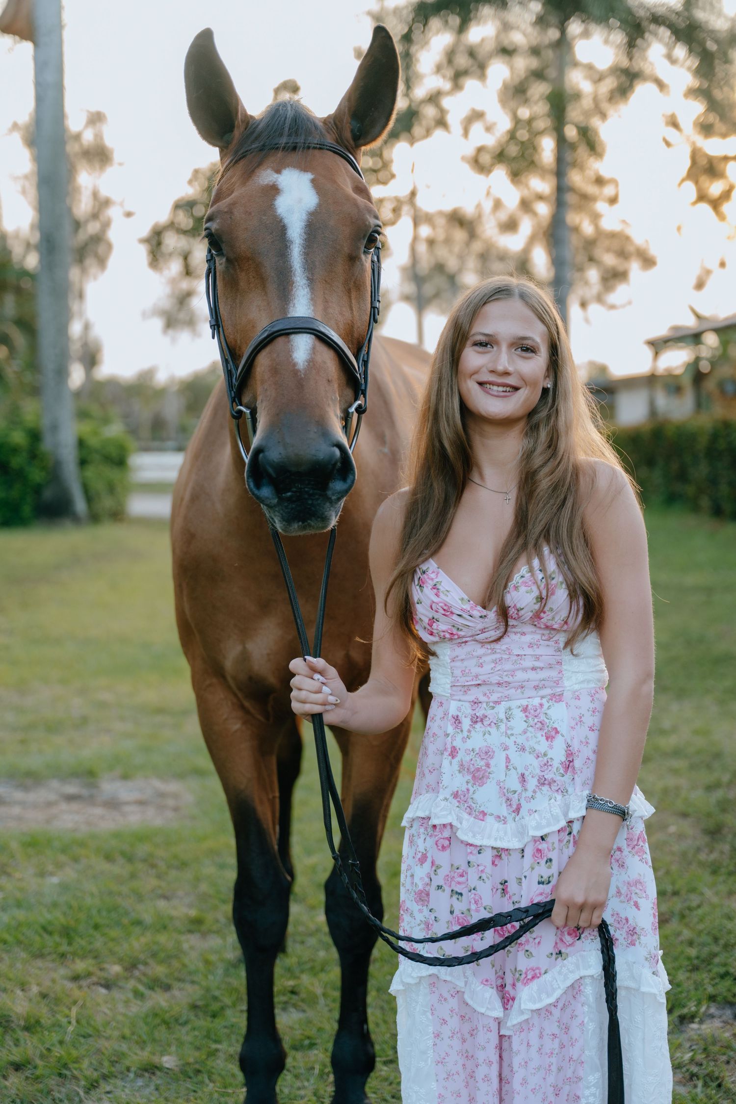 Young woman in floral dress smiling beside a bay horse with white blaze in a sunny outdoor setting.