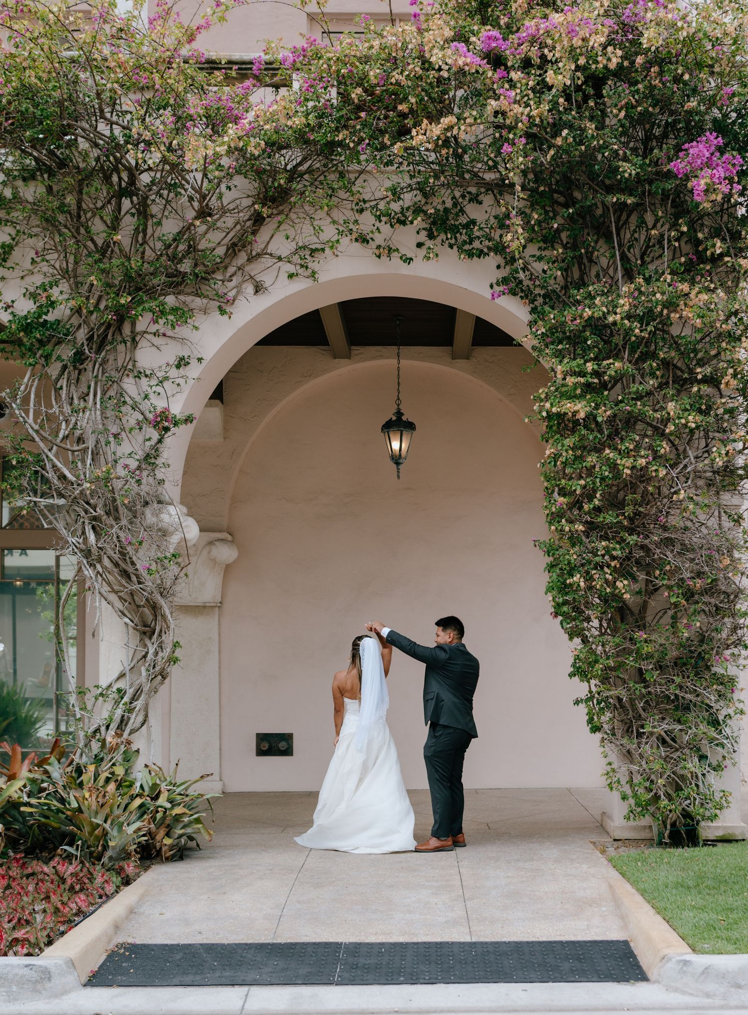 Groom adjusts bride's veil under a flower-covered archway at a romantic outdoor wedding venue.