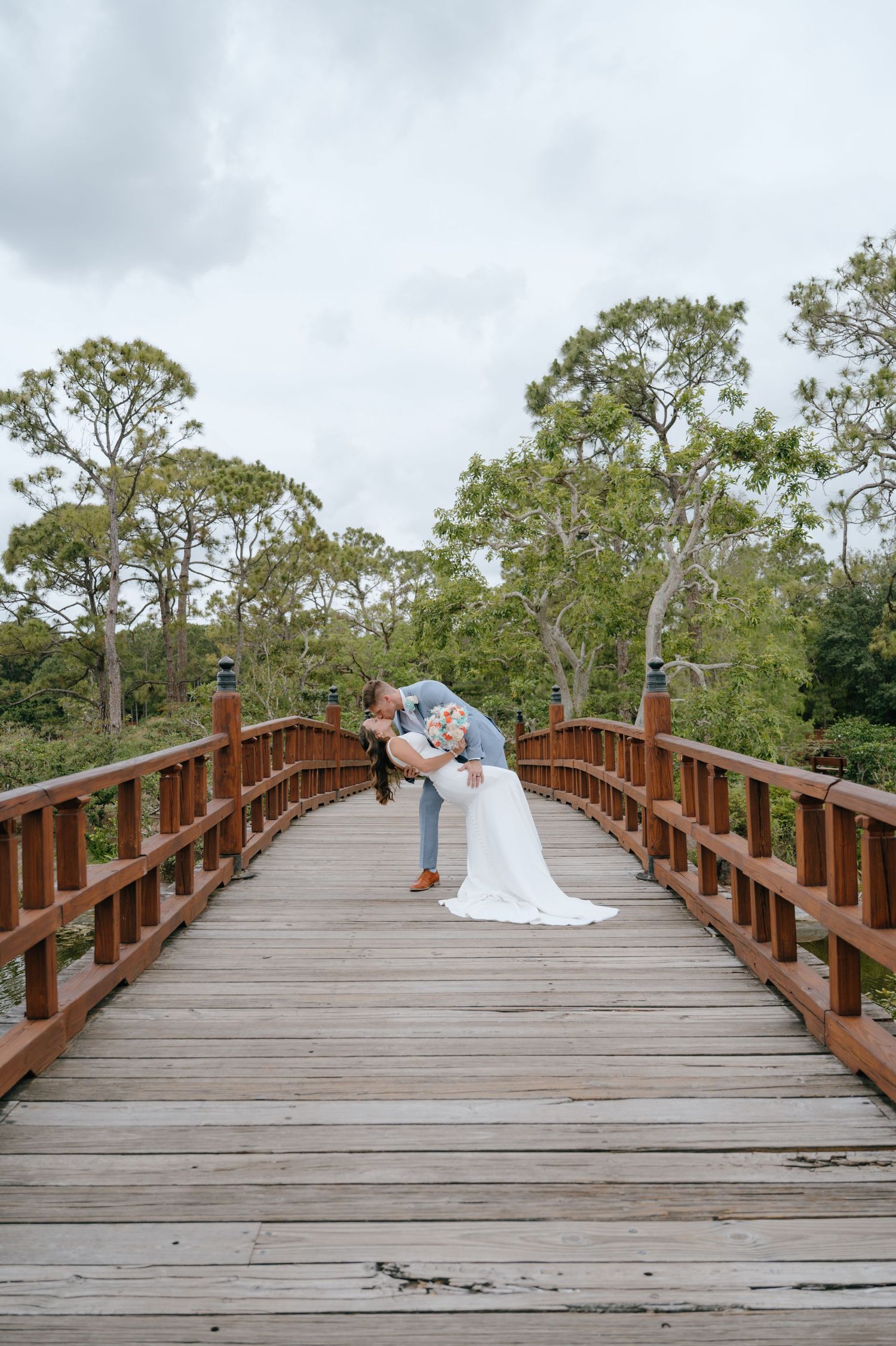 Bride and groom share a romantic dip kiss on a wooden bridge surrounded by lush green trees.