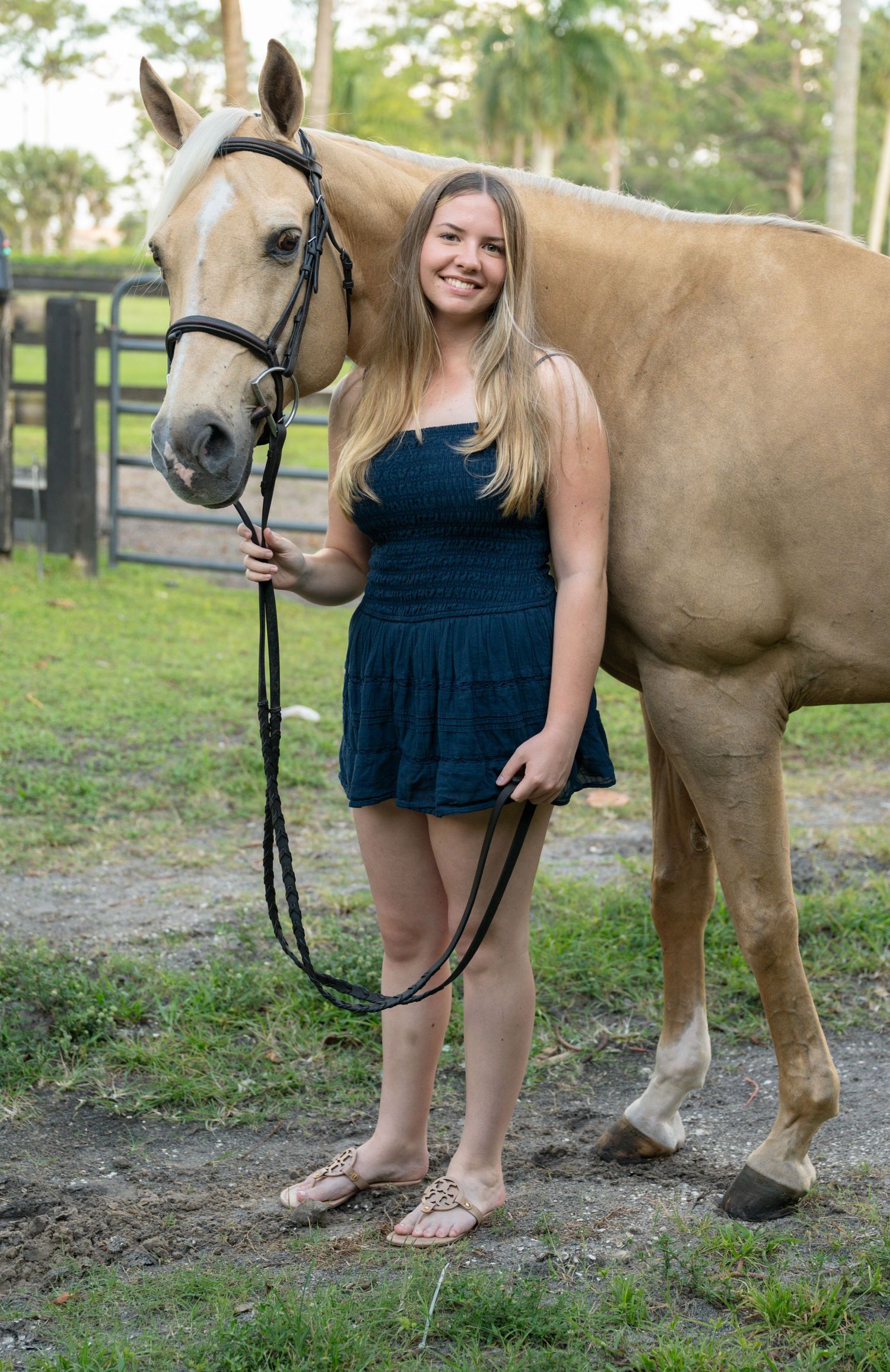 Happy teenage girl in navy dress poses smiling beside palomino horse at sunny equestrian property.