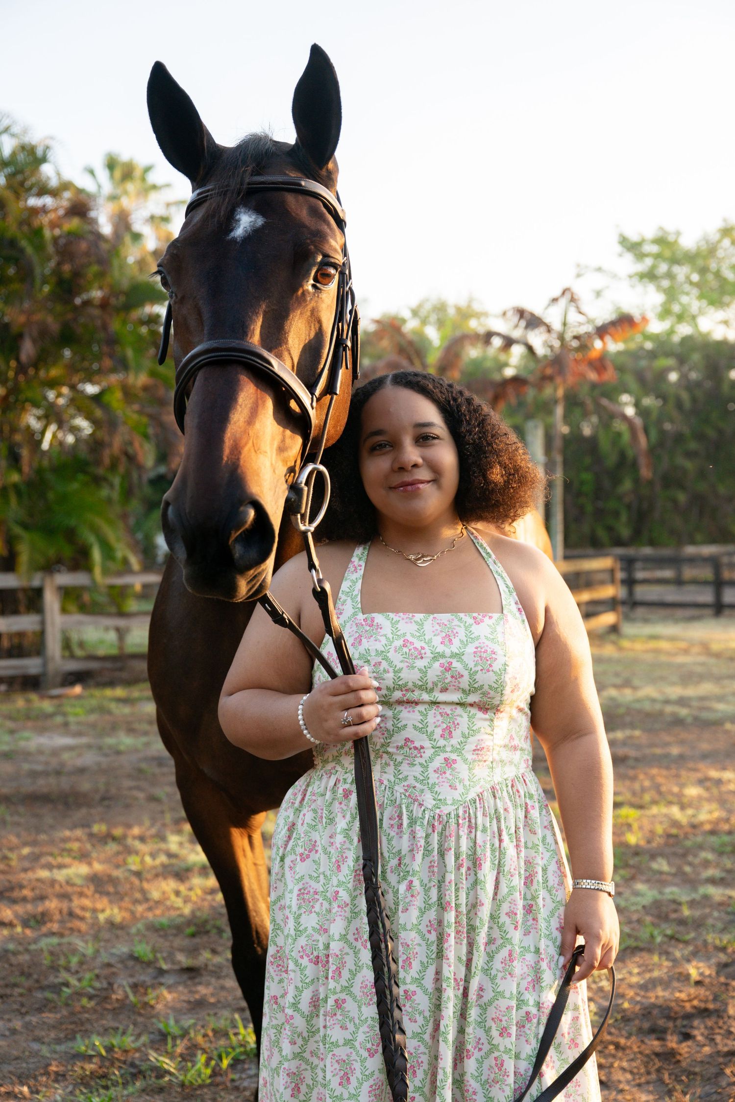 Young woman in floral halter dress poses beside a dark bay horse in an outdoor equestrian setting.
