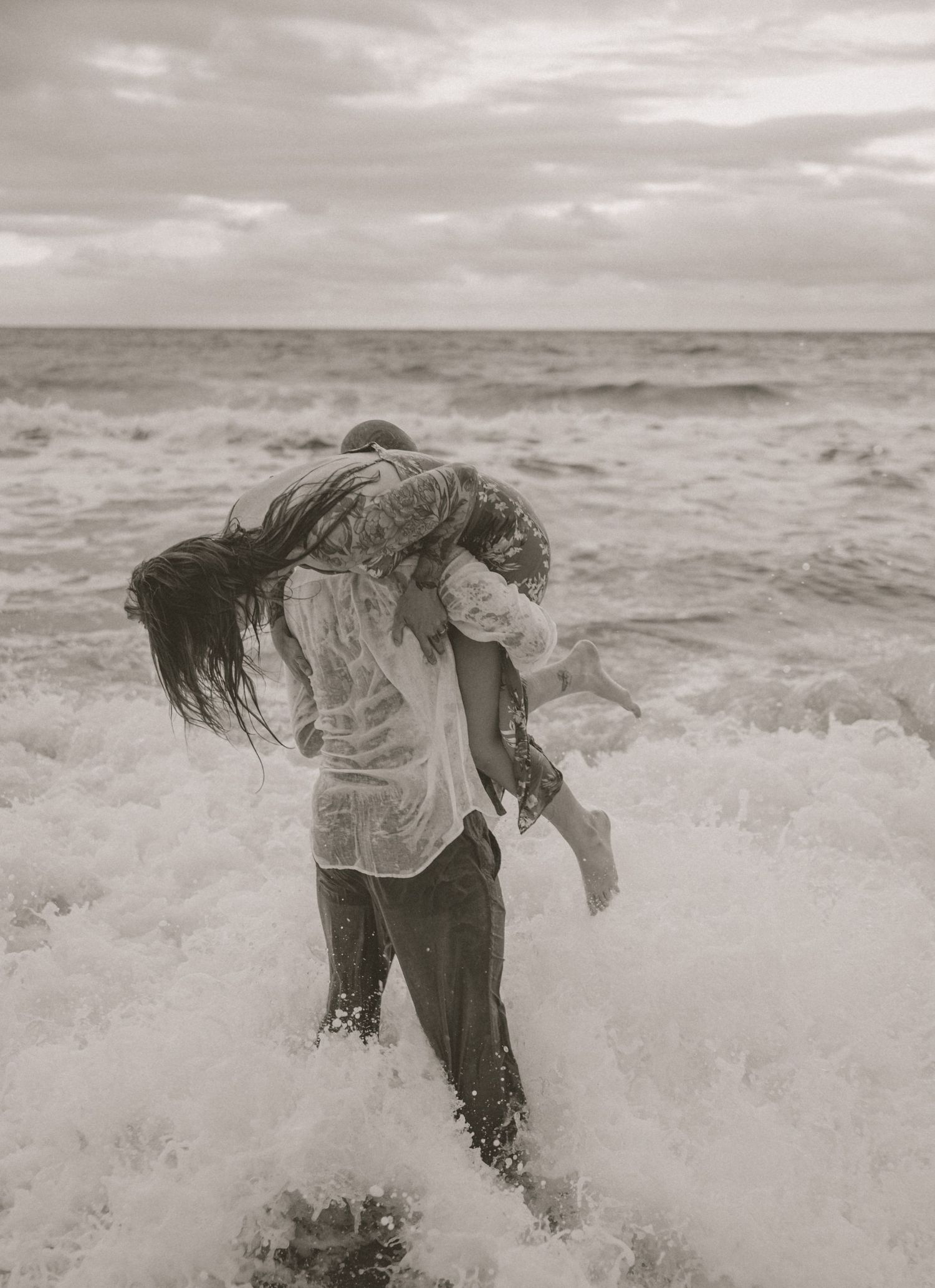 Black and white photo of man carrying woman through crashing ocean waves on stormy beach.