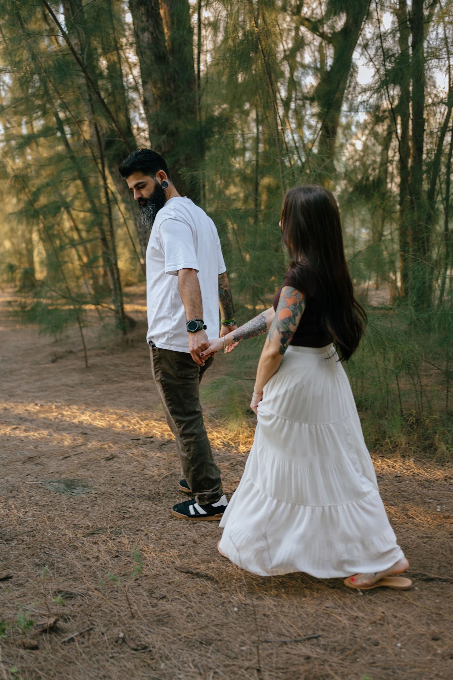 Tattooed couple holding hands walking through a sunlit forest path.