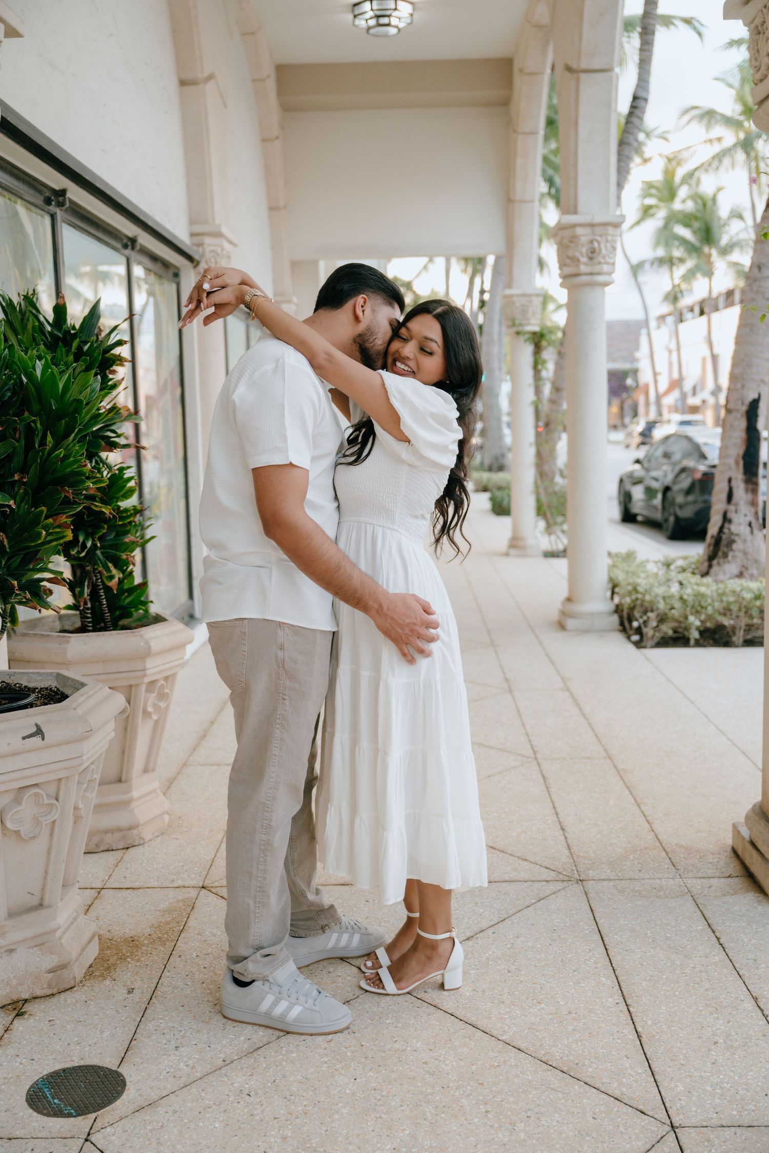 Couple in white outfits embracing and dancing together in a palm tree-lined colonnade.