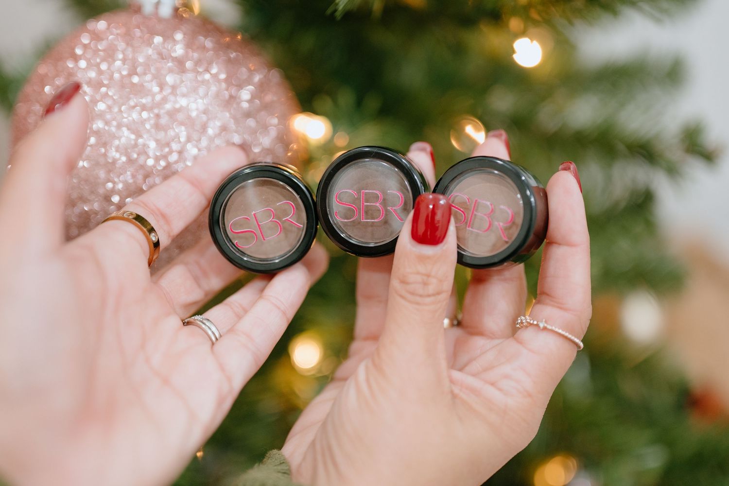 Hands with red nails hold three open circular makeup compacts in front of a glittery holiday Christmas tree.
