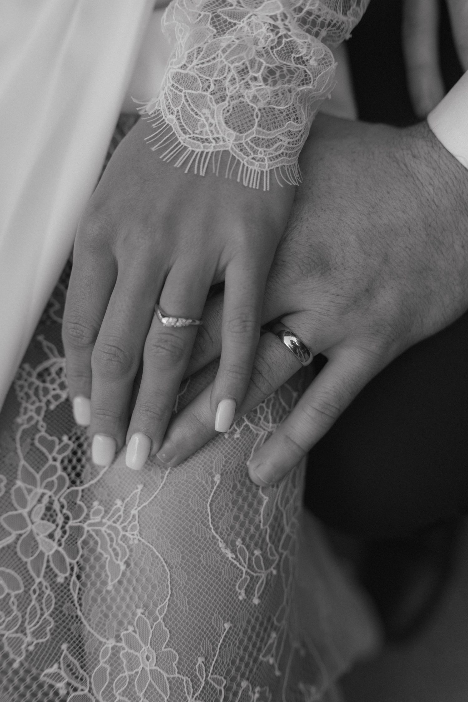 Black and white close-up of bride and groom hands showing wedding rings on lace dress.