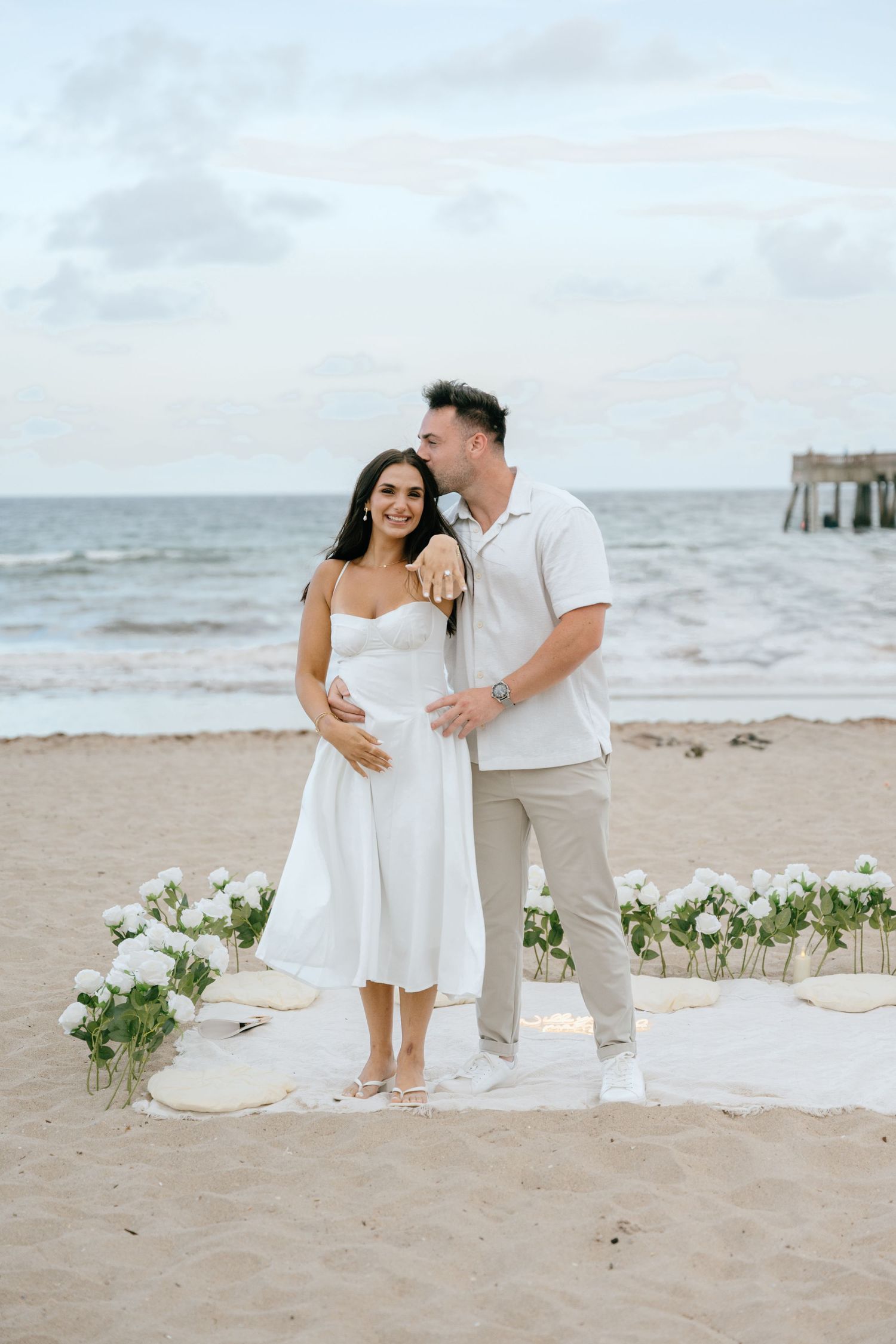 Engaged couple embracing on sandy beach with white floral arrangement and ocean pier in background.