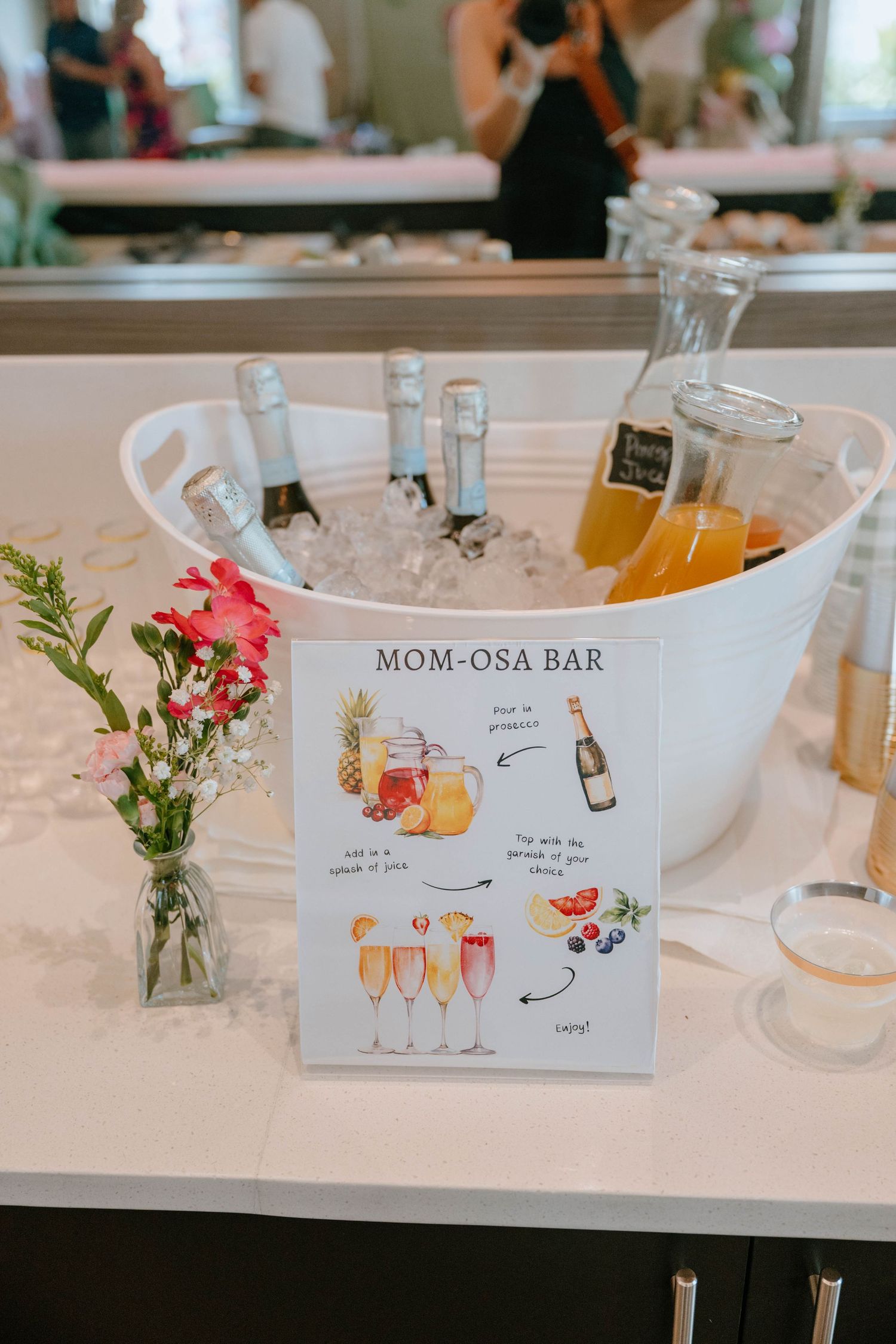 A Mom-osa Bar setup with champagne bottles on ice, juices, and a decorative sign at a baby shower.