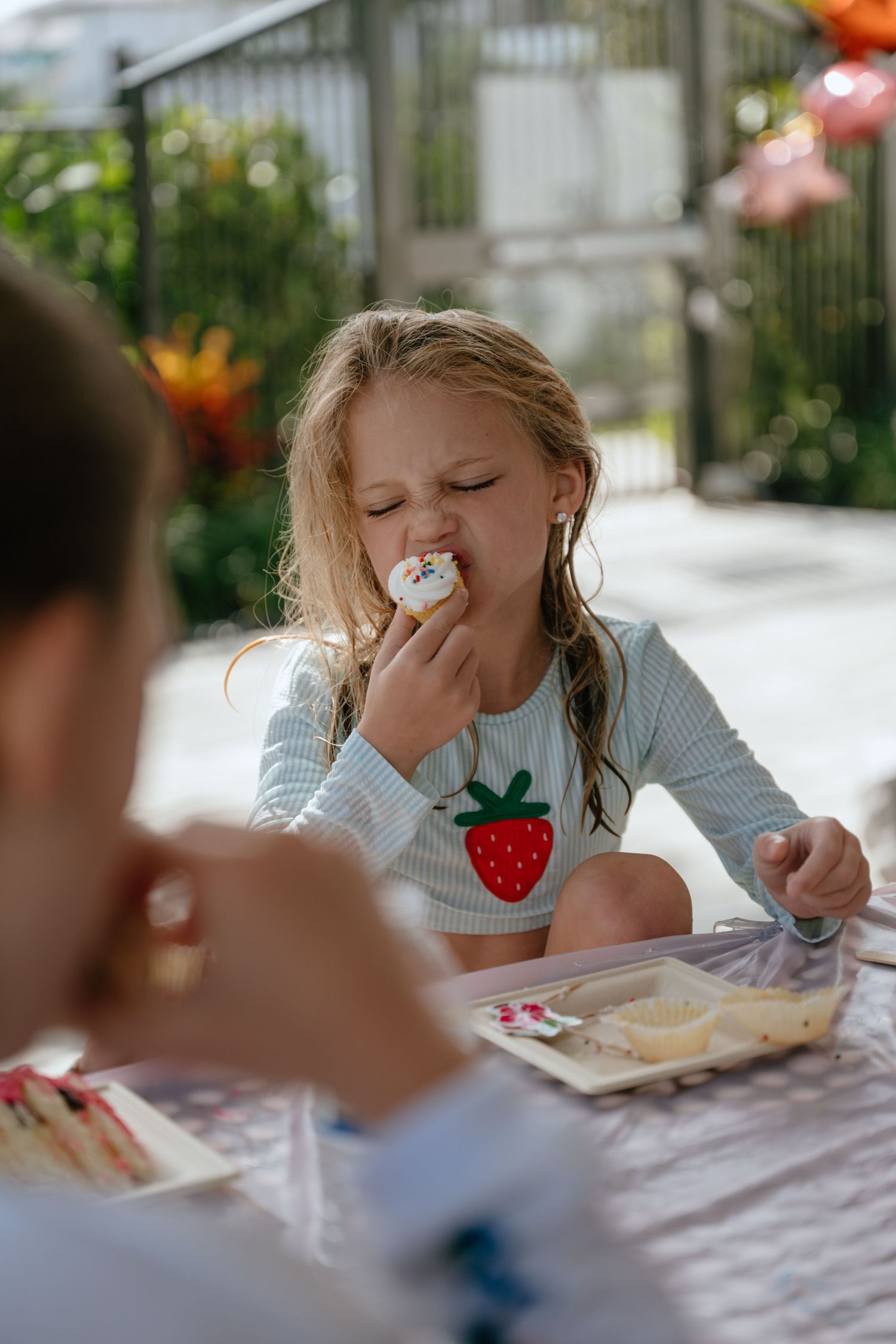 Young girl in strawberry print shirt eating frosting off a cookie at an outdoor birthday party table.