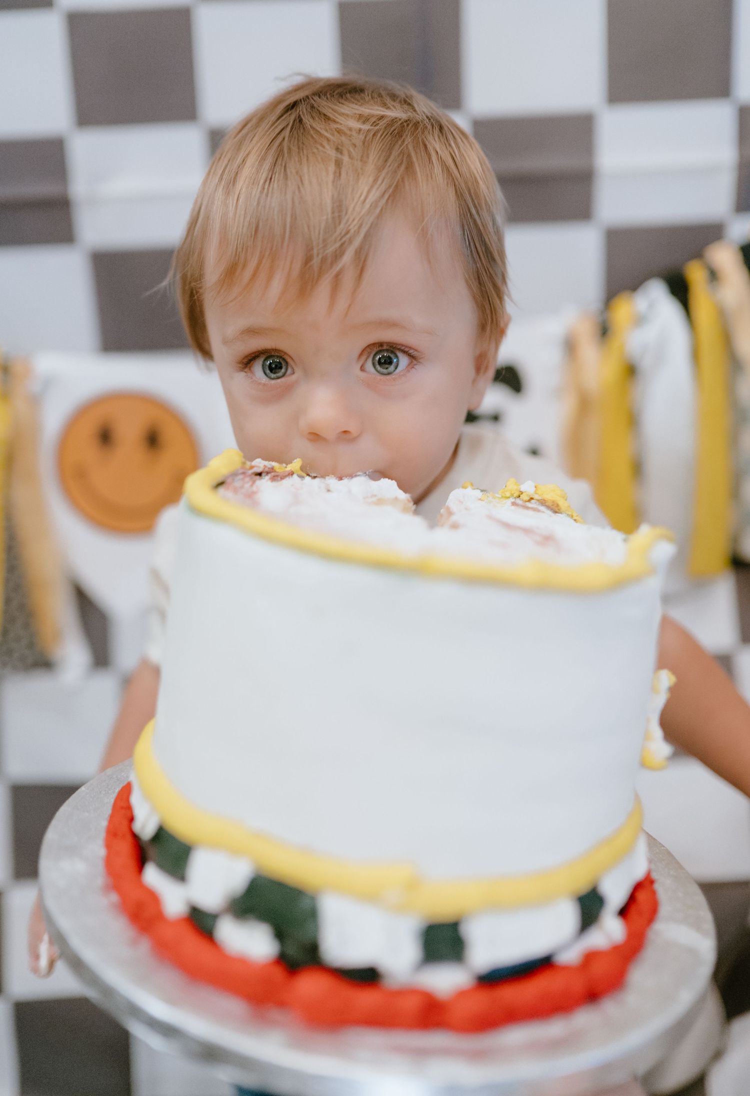 Adorable toddler bites into a decorated first birthday smash cake against a checkered backdrop.