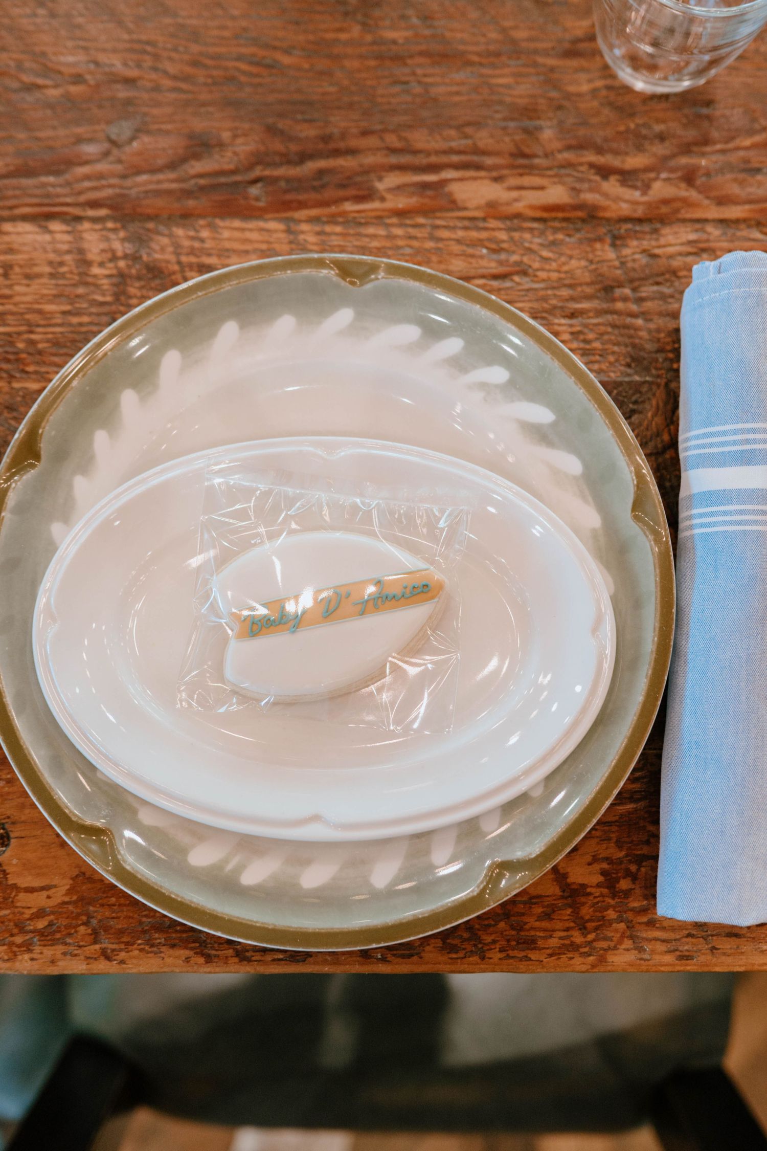 Baby shower place setting with sage green scalloped plate, white dishes, and wrapped cookie favor on wood table.