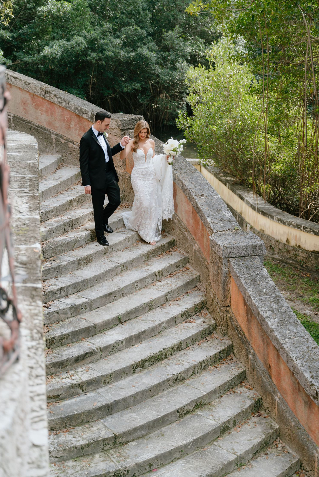 Bride and groom walking hand in hand down curved stone steps at a scenic outdoor wedding venue.