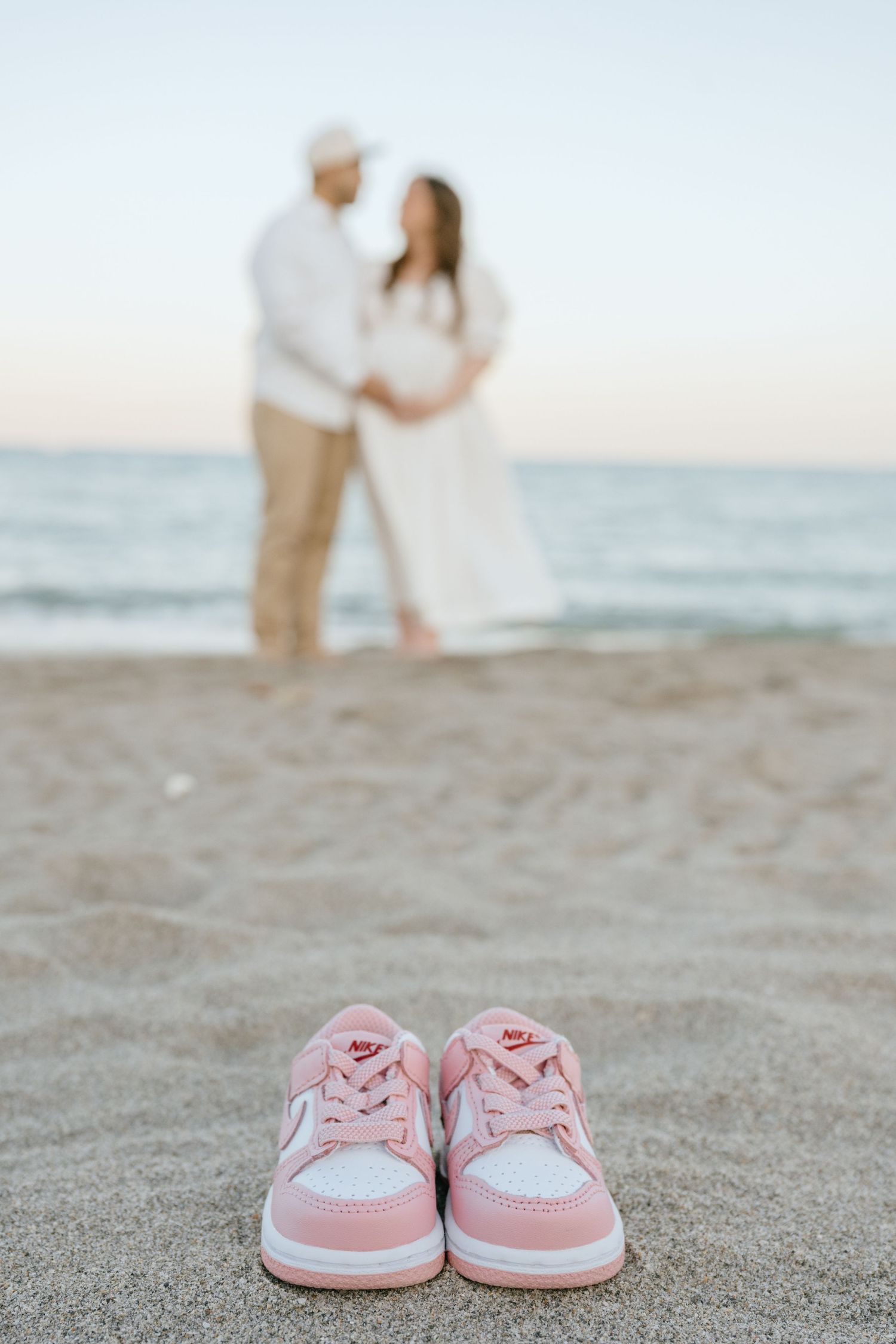 Pink baby Nike sneakers in beach sand with expecting couple embracing at ocean shore in background.