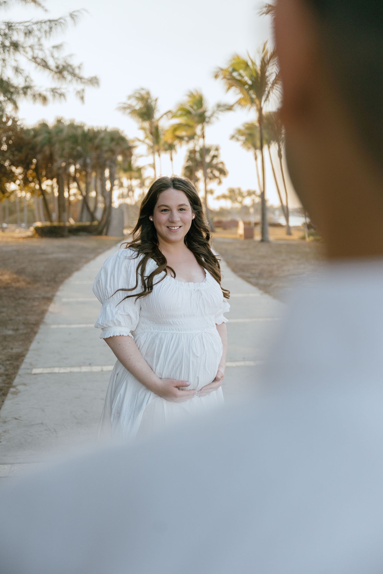 Smiling pregnant woman in white dress holds belly on palm-lined path at golden hour.