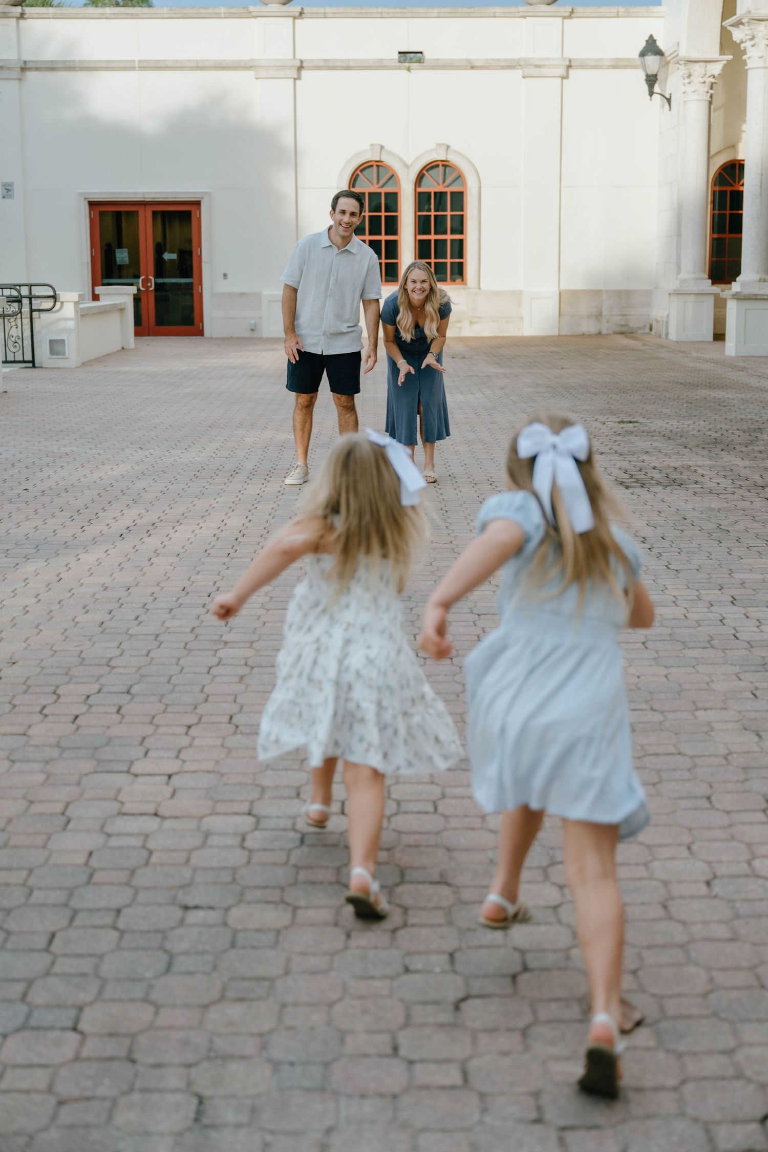 Two young girls in dresses run toward their smiling parents on a sunny brick courtyard.