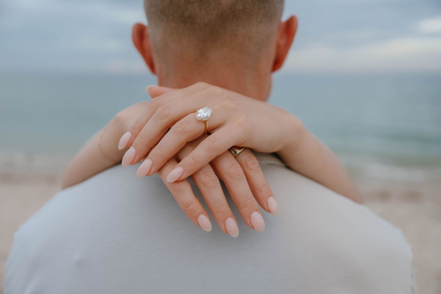Close-up of woman's hand with oval diamond engagement ring resting on man's shoulder at the beach.