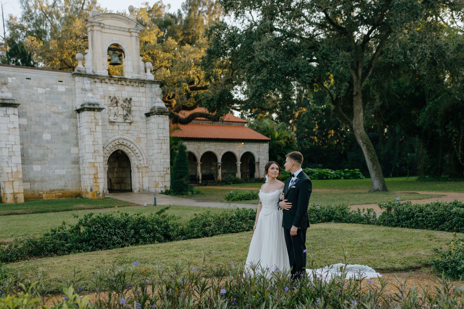 Bride and groom sharing a romantic moment in a formal garden near a historic stone chapel.