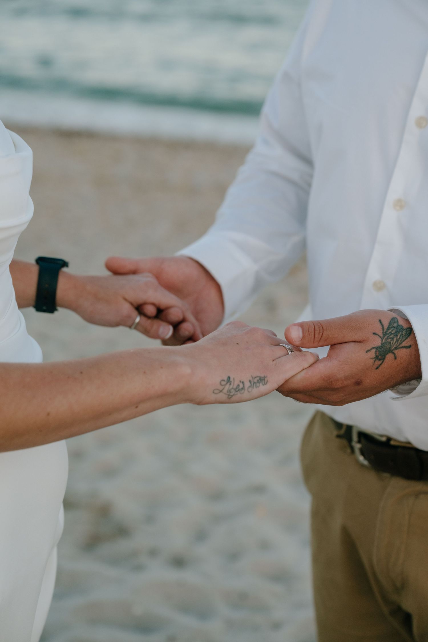 Close-up of bride placing wedding ring on tattooed groom's hand during beach ceremony.