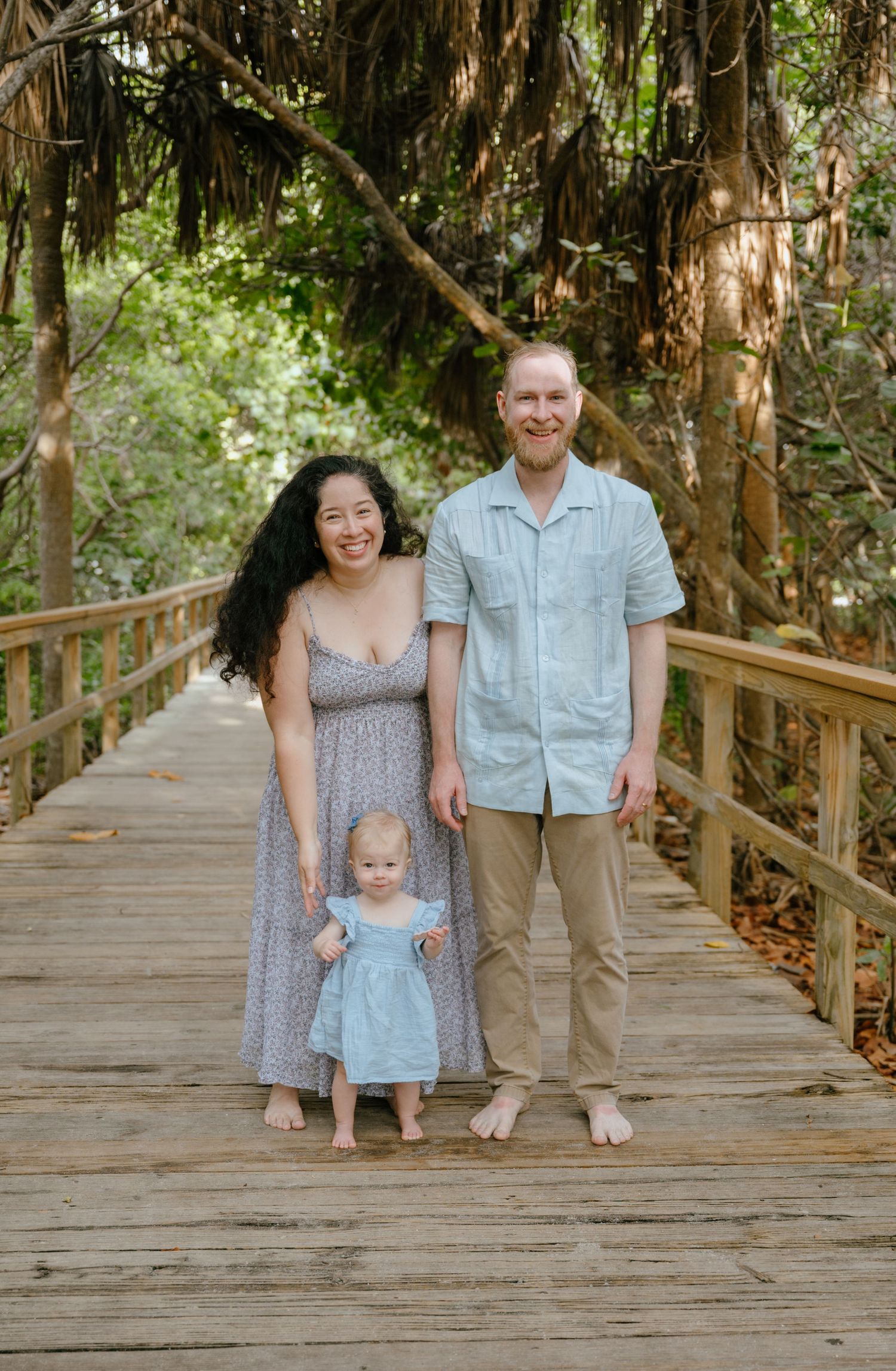 A young family with a baby girl stands barefoot on a wooden boardwalk surrounded by tropical trees.