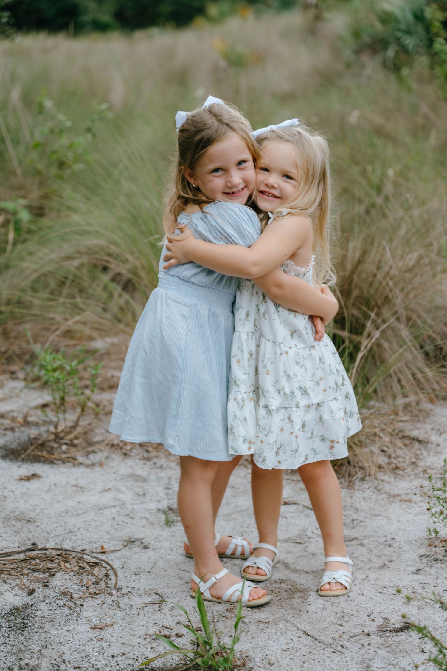 Two little girls in matching white bows and pastel dresses hug each other on a sandy outdoor path.