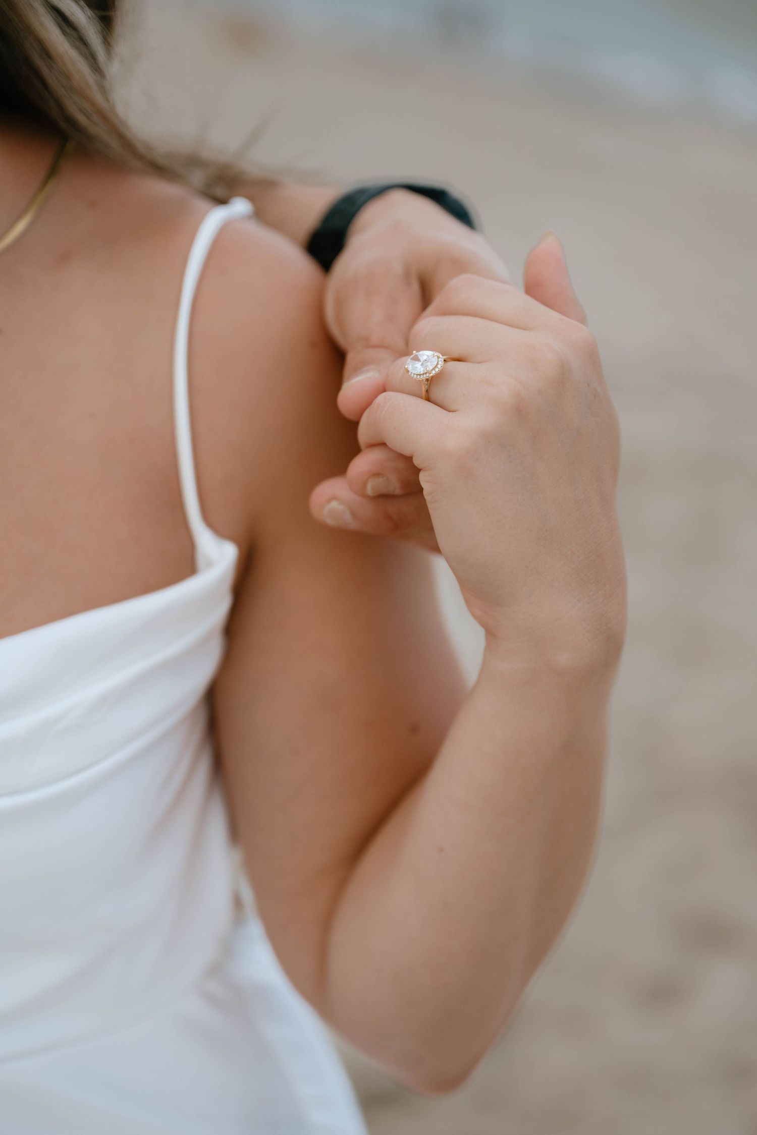 Detail shot of woman's hand displaying an oval solitaire engagement ring while holding her white dress strap.