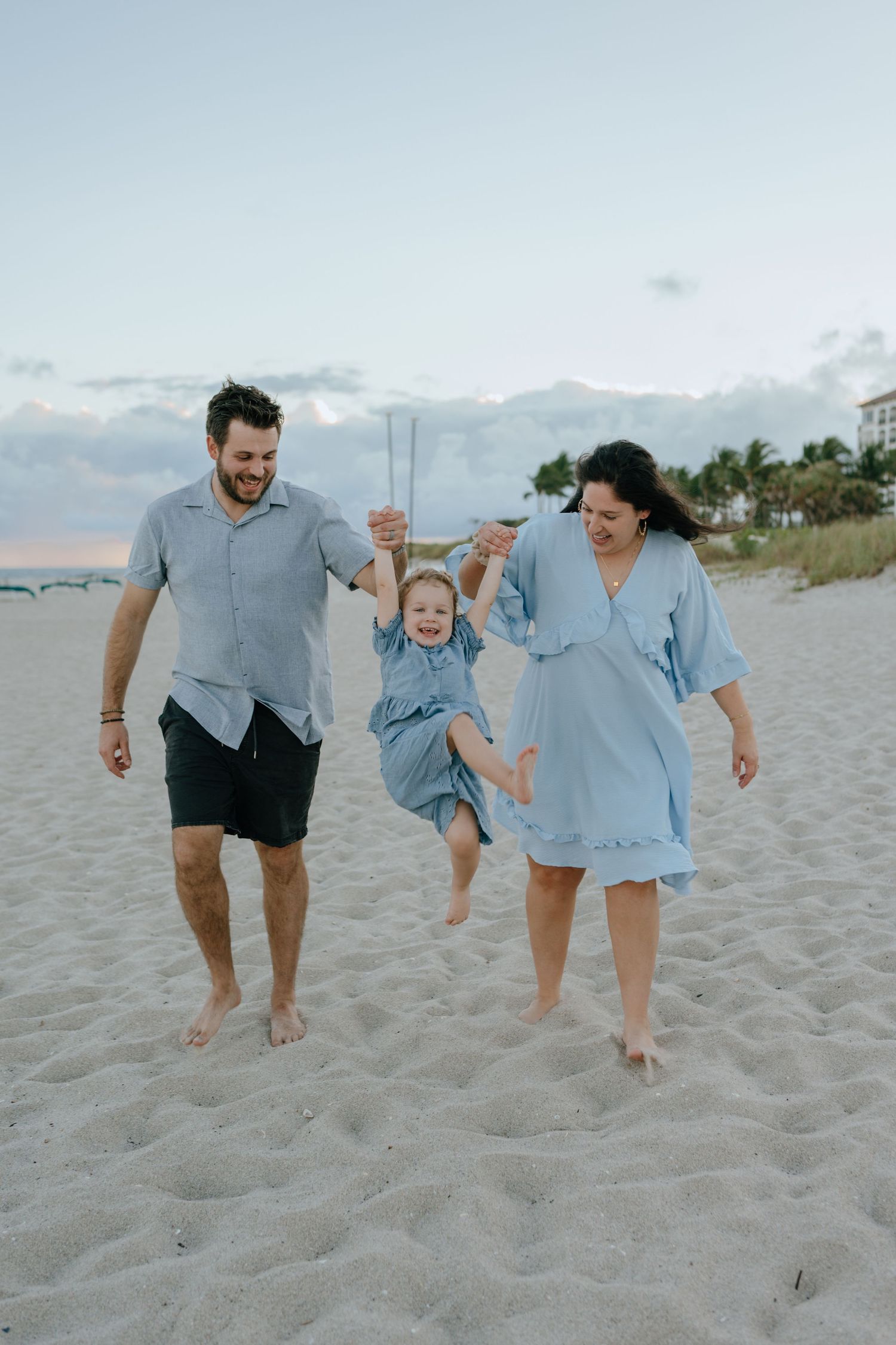 Happy young family swinging their toddler daughter between them while walking barefoot on a sandy beach at sunset.