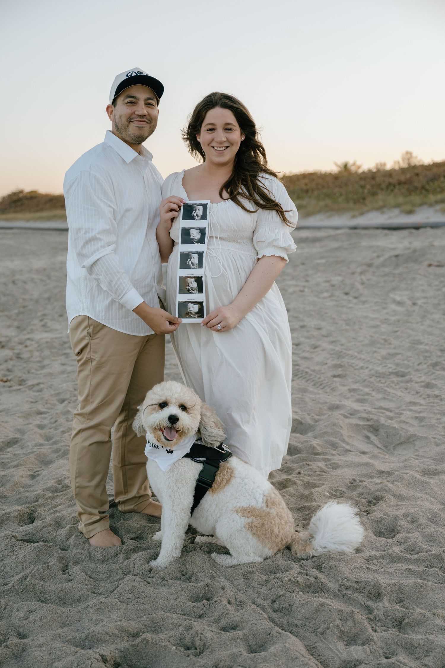 Smiling pregnant couple holding ultrasound strip photos on beach with fluffy white dog at sunset.