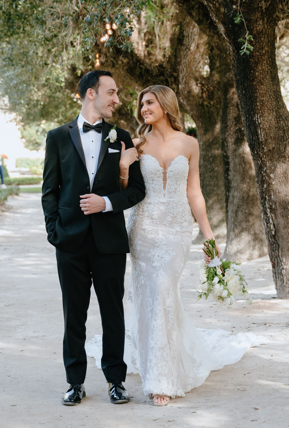 Smiling bride and groom walking together under oak trees, bride wearing a lace fitted gown with bouquet.