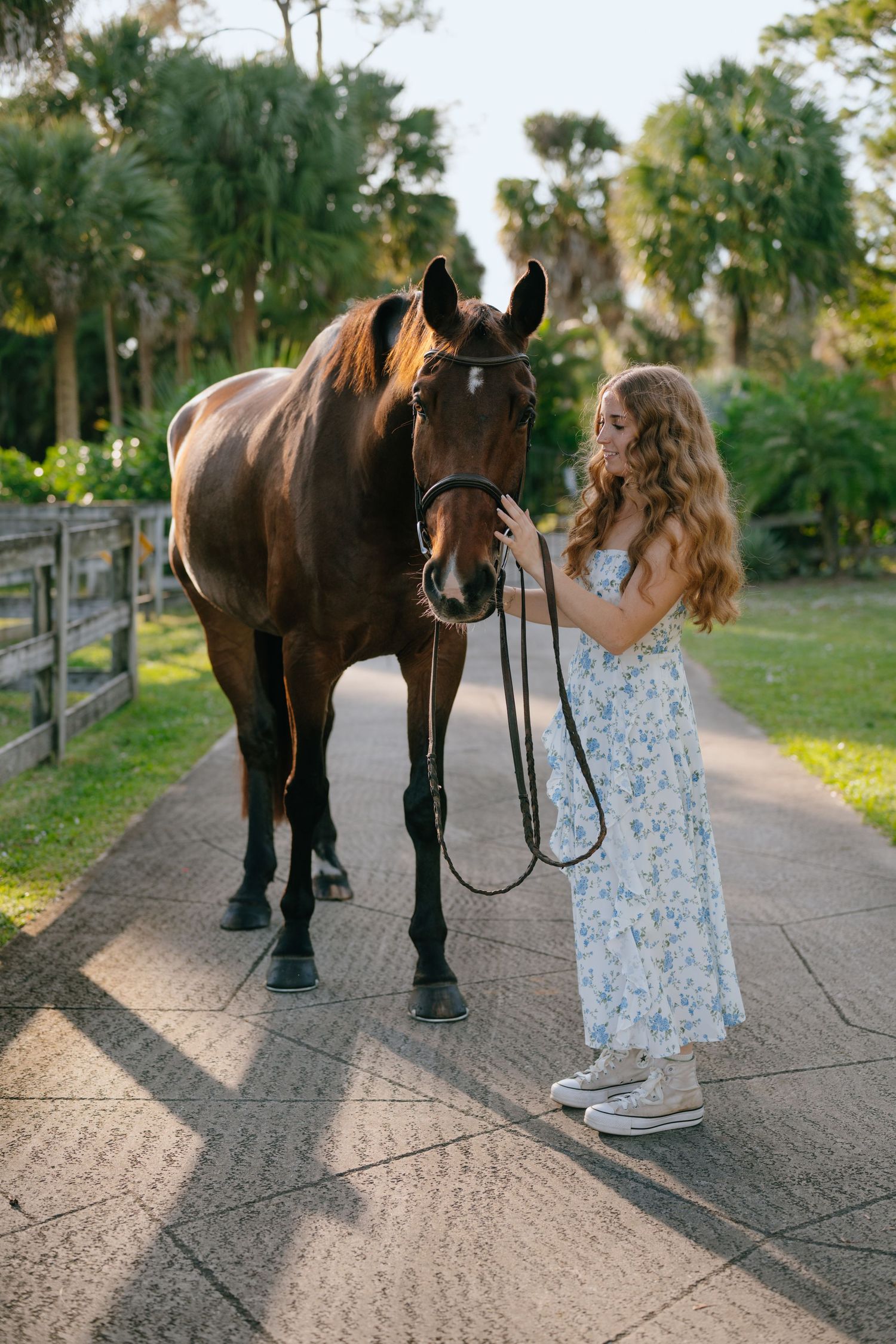 Girl in blue floral dress tenderly pets dark bay horse on sunlit equestrian path.