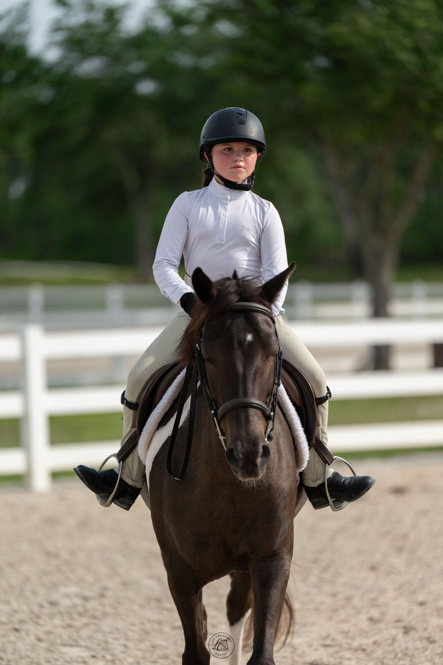 Young equestrian in black helmet riding dark horse toward camera in outdoor riding arena.