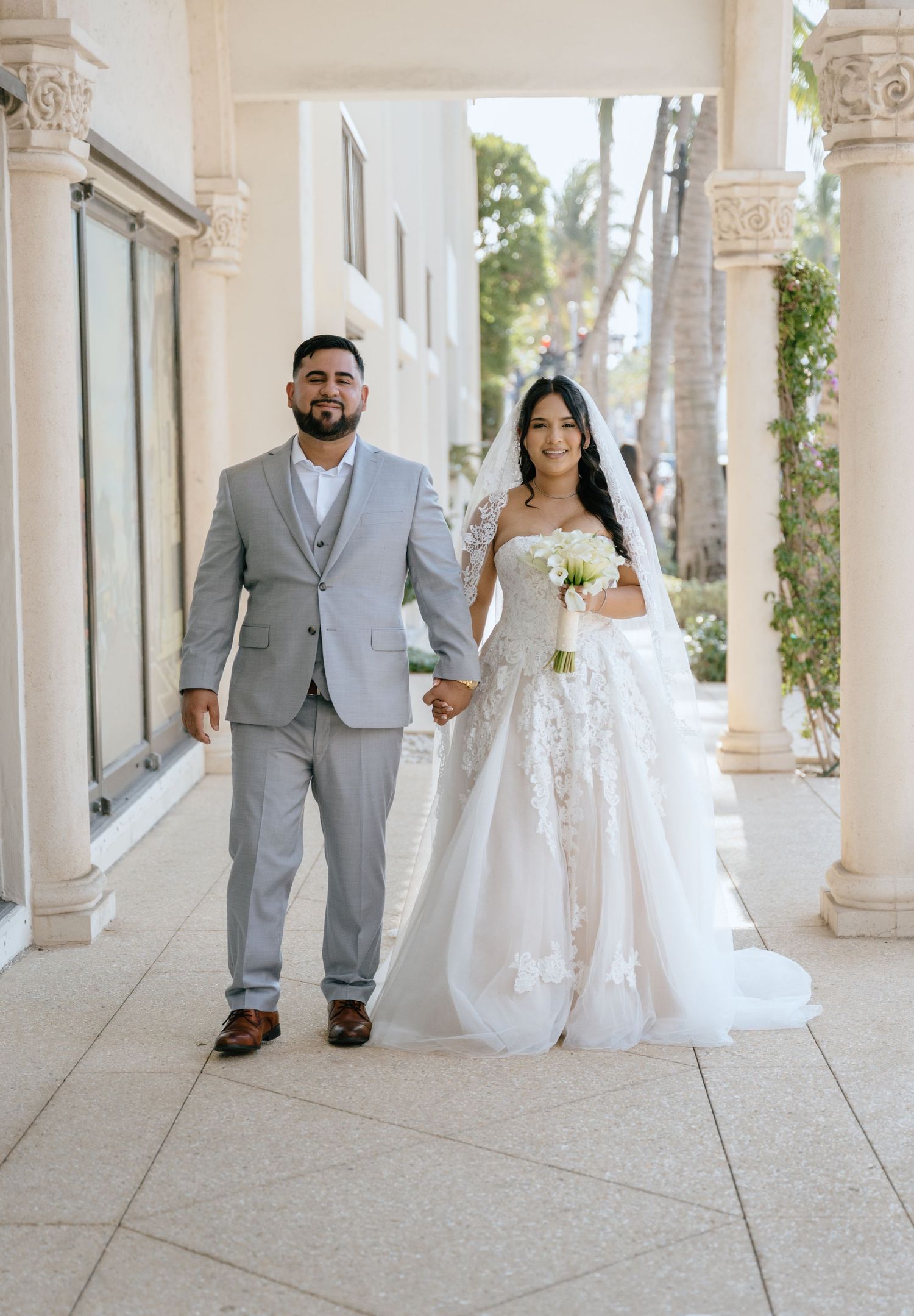 Bride in lace ball gown and groom in gray suit walk hand in hand at outdoor venue.