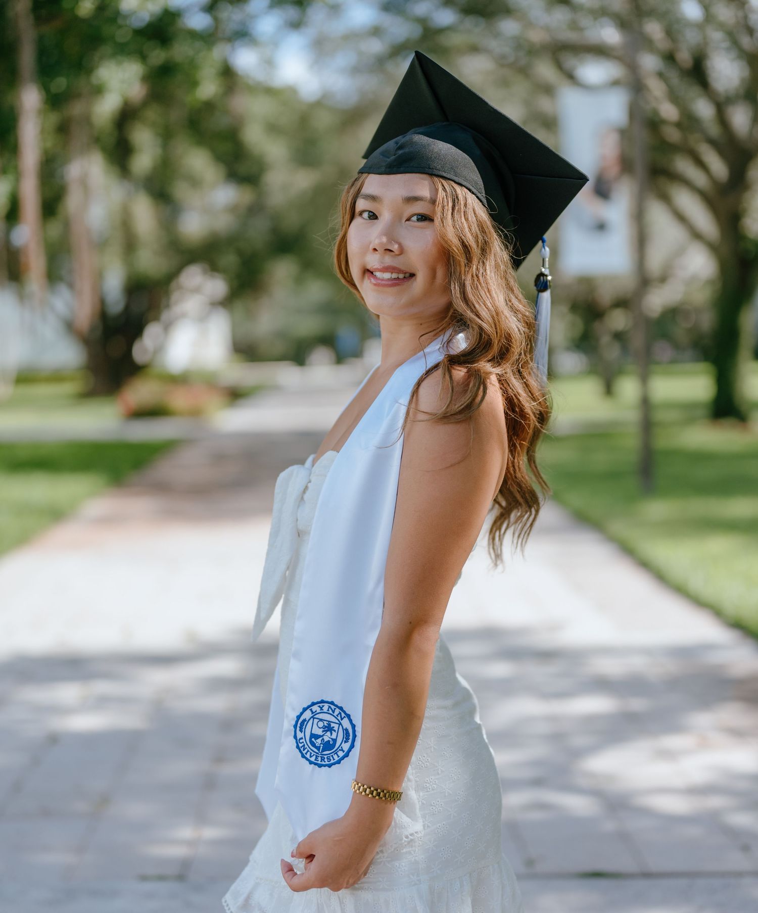 Smiling graduate in white dress and black cap holding stole with Lynn University seal outdoors.