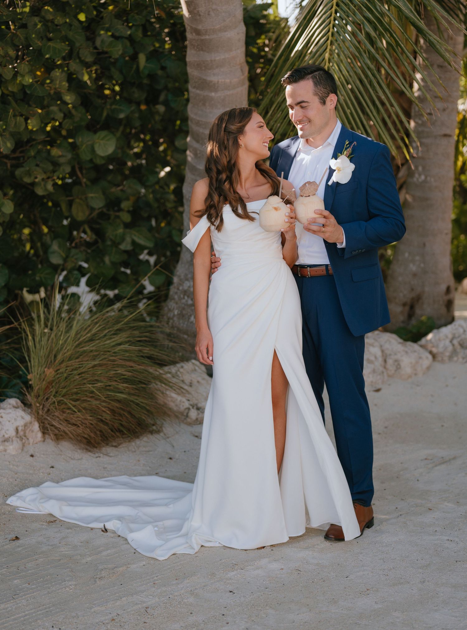 Bride and groom smiling together in wedding attire among tropical palm trees at a beach venue.