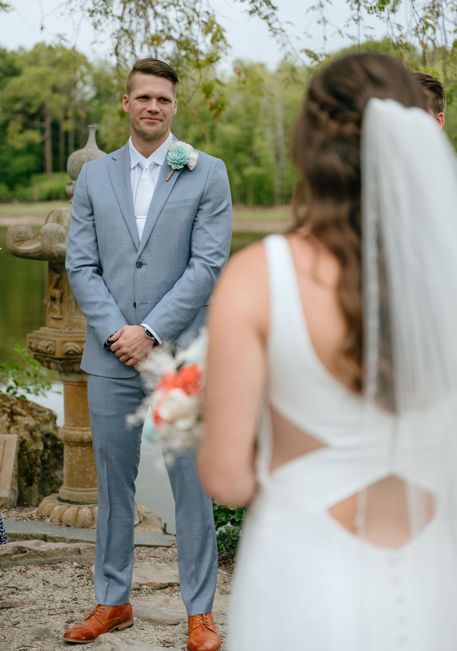 Emotional groom in light blue suit sees bride for the first time during outdoor first look near pond.