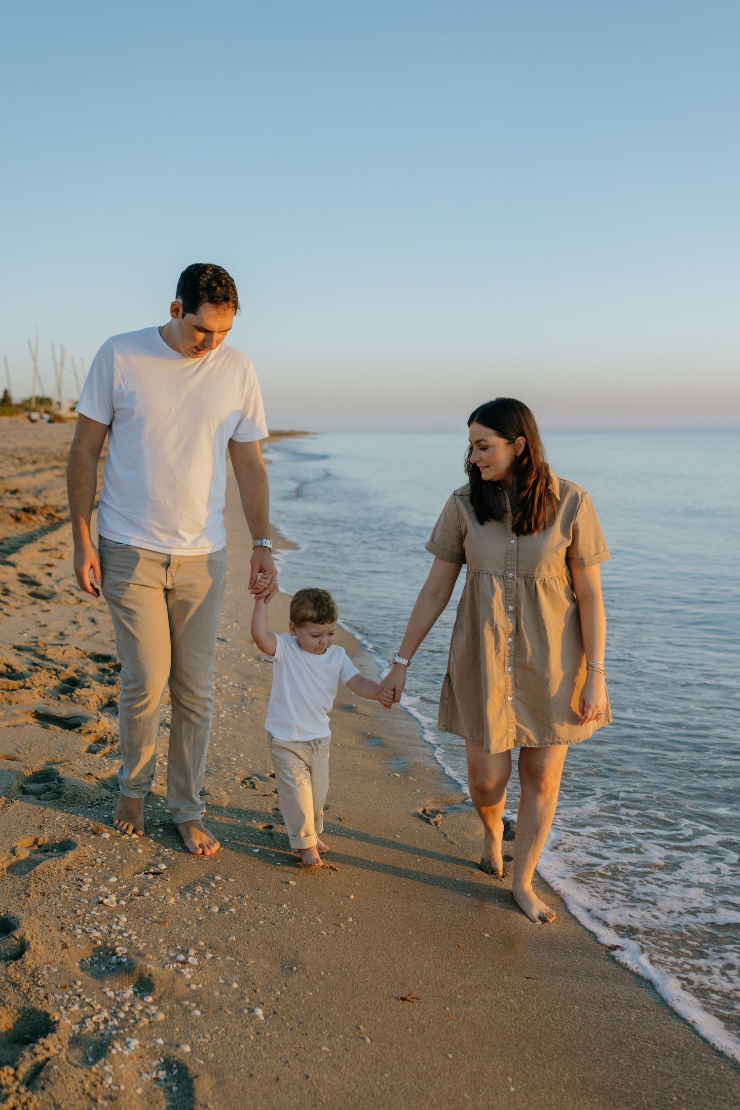 A family of three walks hand-in-hand along a sunny beach at golden hour.